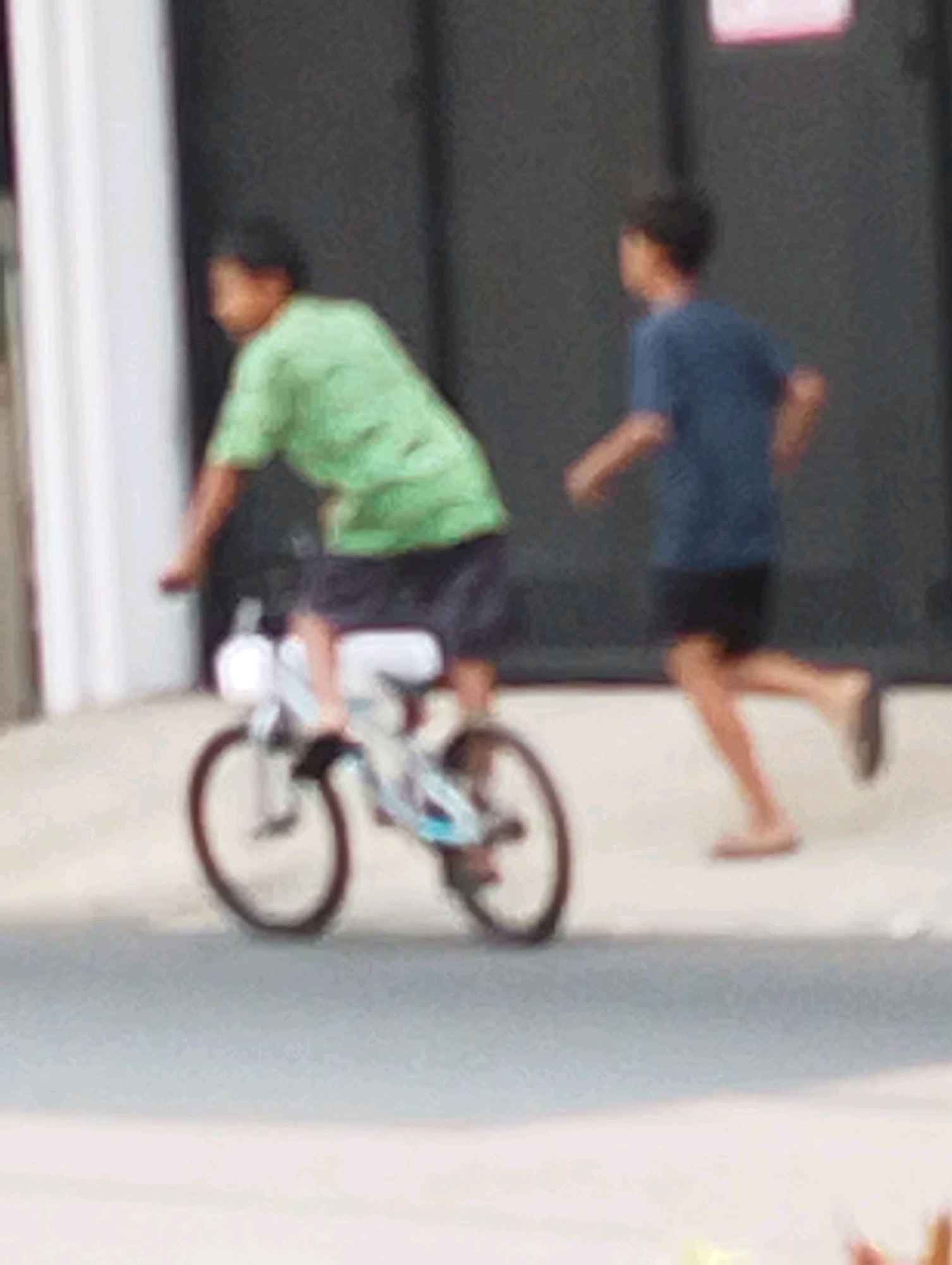 In Gempeng, Indonesia, two boys are captured in a low-resolution outdoor scene. On the left, a boy in a green t-shirt rides a white bicycle, with dark shorts. The bicycle has visible spokes and a white seat. To the right, another boy in a blue t-shirt and dark shorts is running, with one leg extended forward. The background appears to be a dark wall, possibly a building, with a hint of a light-colored vertical structure on the far left. The ground is a light-colored pavement or road.