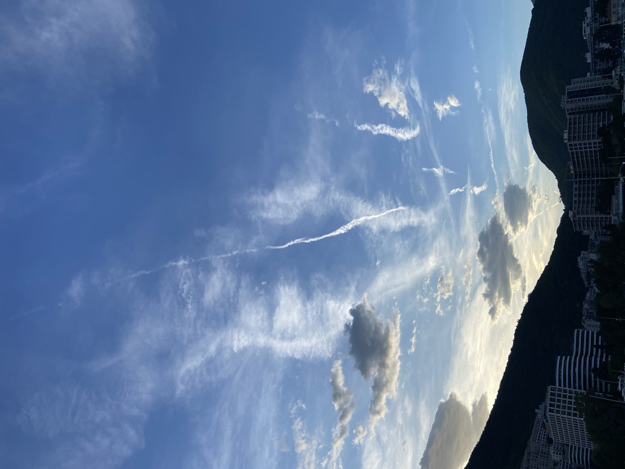The image depicts a partially cloudy sky over a city. Streaks of clouds and contrails crisscross the bright blue sky. Along the right edge of the image is a dark, heavily-forested mountain with building structures along its lower slopes. The location is Unknown, Unknown.