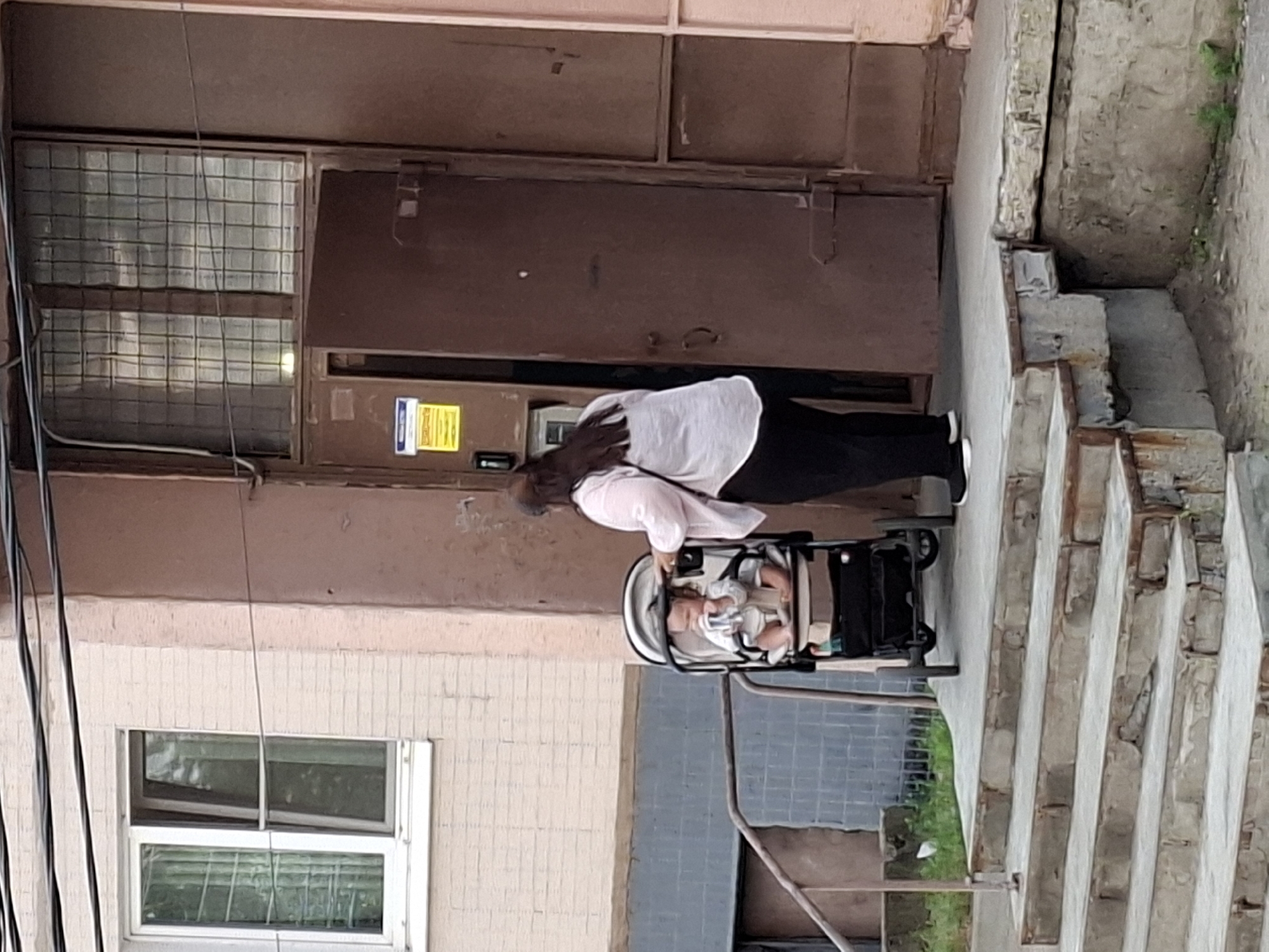 An adult, likely female, with dark hair, wearing a light-colored long-sleeved shirt and black pants, stands to the right of a baby stroller. The stroller contains a young child, seated and facing forward, who appears to be drinking from a bottle. The adult is positioned behind the stroller, with their right hand on its handle, suggesting they are pushing or about to push it.
The scene is set outdoors, in front of a building with a reddish-brown metal door. The building facade appears to be constructed with light-colored bricks, painted a pale pink. To the left of the door, there is a window with a white frame and a metal security grate. A set of concrete stairs leads up to the entrance. The ground is paved, and some grass and vegetation are visible around the base of the building and stairs. The overall environment suggests a residential building entrance in an urban setting. The image is taken from a slightly low angle.