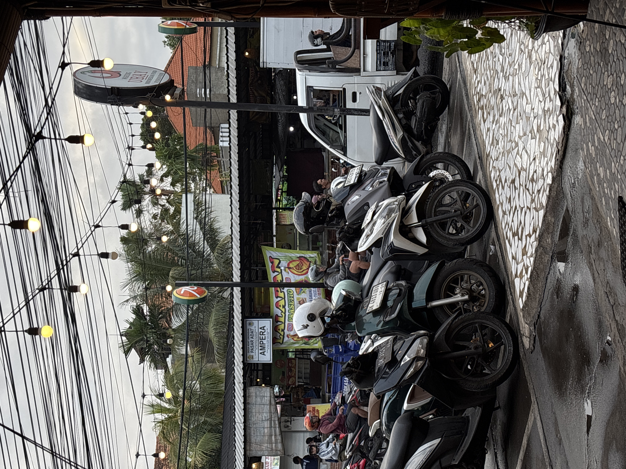 The image depicts a street scene in Kuta Selatan, Indonesia, likely an outdoor dining area or a street-side establishment. In the foreground, a row of parked motorbikes is visible, with several individuals seated on them, some wearing helmets. The motorbikes are of various colors and models, and some have license plates clearly visible.
Behind the motorbikes, a white pickup truck is parked, with two individuals inside. One person is in the driver's seat, and another is in the passenger seat. A sign reading "PASAR ADAT AMPERA" is mounted on a pole, suggesting a local market or community area. Adjacent to this sign, a large banner with text and images of chickens indicates a food vendor, likely selling Indonesian cuisine.
Overhead, a dense network of power lines crisscrosses the sky, strung with numerous light bulbs, many of which are illuminated, casting a warm glow. This suggests the image was taken in the late afternoon or early evening. Tropical palm trees and reddish-tiled roofs of buildings are visible in the background, contributing to the tropical ambiance of the location. The ground is a mix of wet asphalt and a mosaic-tiled pattern, with puddles of water indicating recent rain. The overall scene suggests a busy, somewhat chaotic urban environment typical of parts of Kuta Selatan.