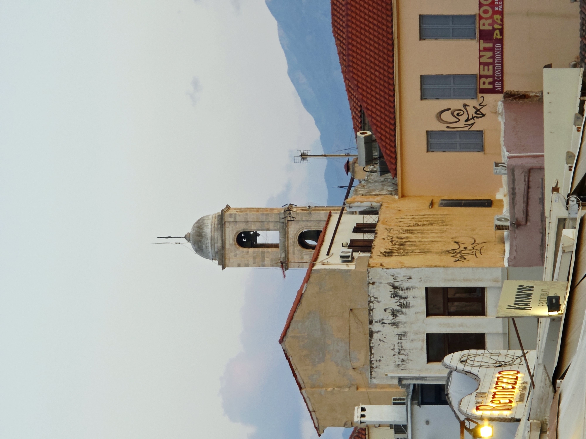 The image shows a view of buildings in the old town of Chania, Greece, with a bell tower of a church in the background. The foreground features several buildings with red tiled roofs and weathered facades. Signs for "Remezzo" and "Kavouras Restaurant" are visible, indicating commercial establishments. Air conditioning units are mounted on some of the buildings. In the distance, mountains are visible against a pale sky with scattered clouds. The overall impression is of a warm, sunny day in a historic Mediterranean city.