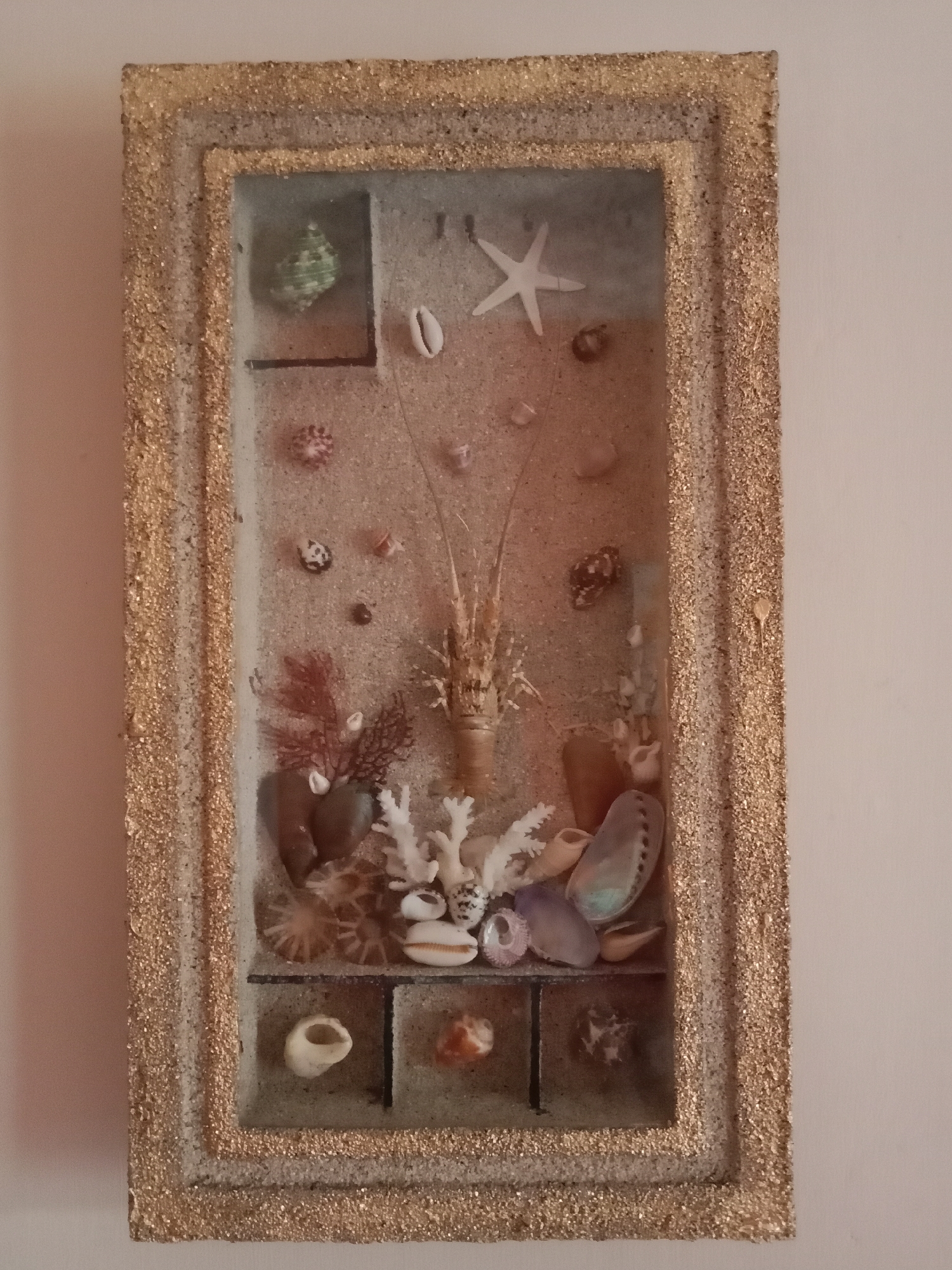 The image shows a framed diorama of seashells and other beach-related items. The diorama is rectangular and appears to be made of glass with a sandy interior. It contains various shells, including a starfish, conch shells, and smaller, spiral shells. There are also some pieces of coral and what appears to be a dried crustacean or shrimp in the center. The frame itself is textured with what looks like glitter or small particles, giving it a golden hue. The diorama is hanging on a light-colored wall. There is no indication of the location being in Kutosari, Indonesia.