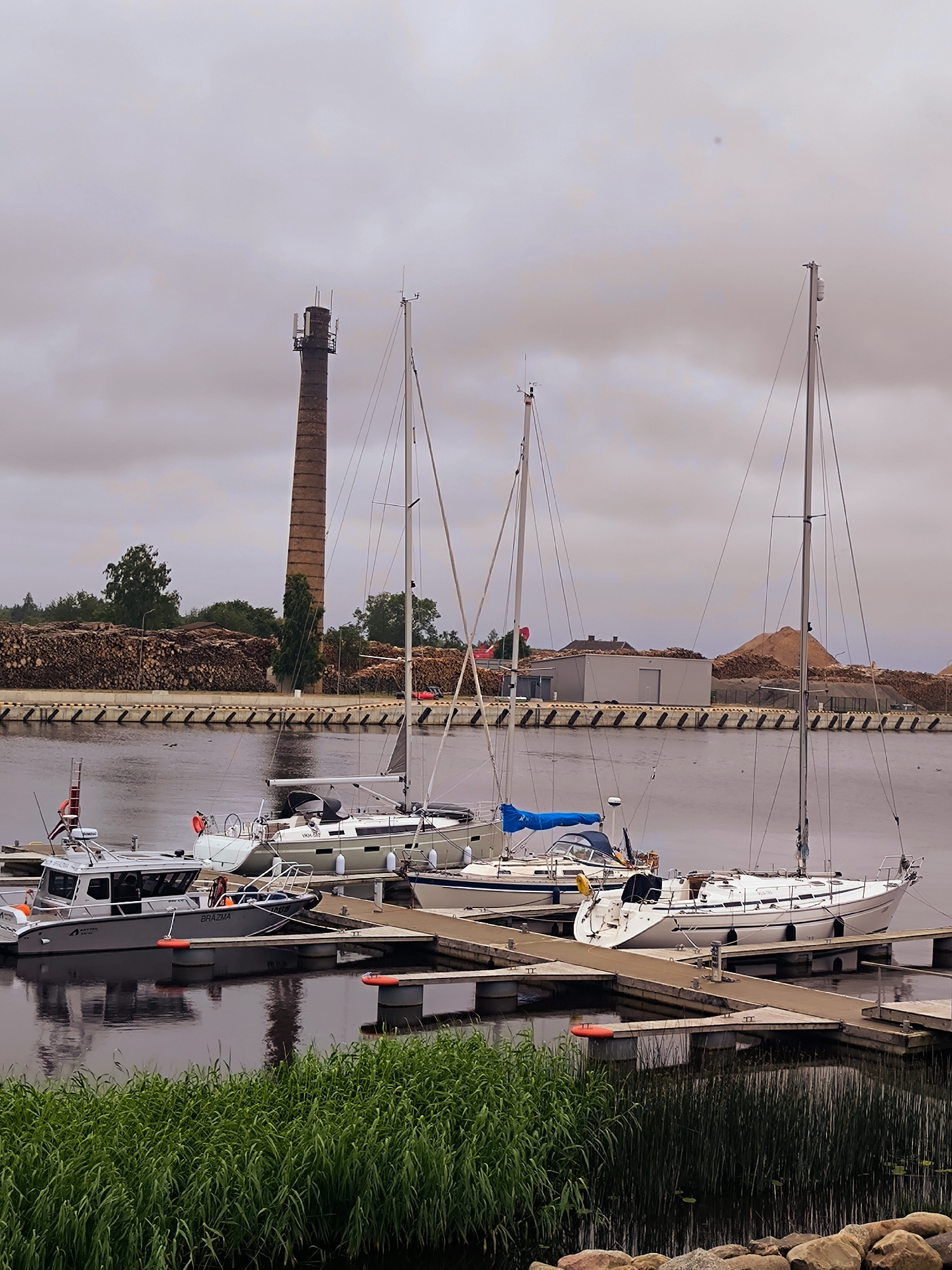 The image depicts a harbor scene in Salacgrīva, Latvia, under an overcast sky. In the foreground, lush green reeds and large rocks line the edge of the water. Several boats are moored at a dock.
The most prominent boats include two sailboats and a smaller motorboat. One sailboat, a white hull with blue trim, is positioned furthest to the right, its mast reaching high into the sky. Another sailboat, also white with blue accents, is docked to its left, with a blue sail cover visible. A grey motorboat, labeled "BRAZMA," is docked to the left of the sailboats, featuring a cabin and a ladder on its side.
In the background, a tall, weathered brick chimney stands out against the sky. Behind the boats and the chimney, a large expanse of stacked timber logs is visible, suggesting a nearby lumber processing facility or storage area. A simple, light-colored building is situated amongst the timber piles. The water is calm, reflecting the boats and the sky. The overall atmosphere is one of a functional port or marina, with a mix of leisure and industrial elements.