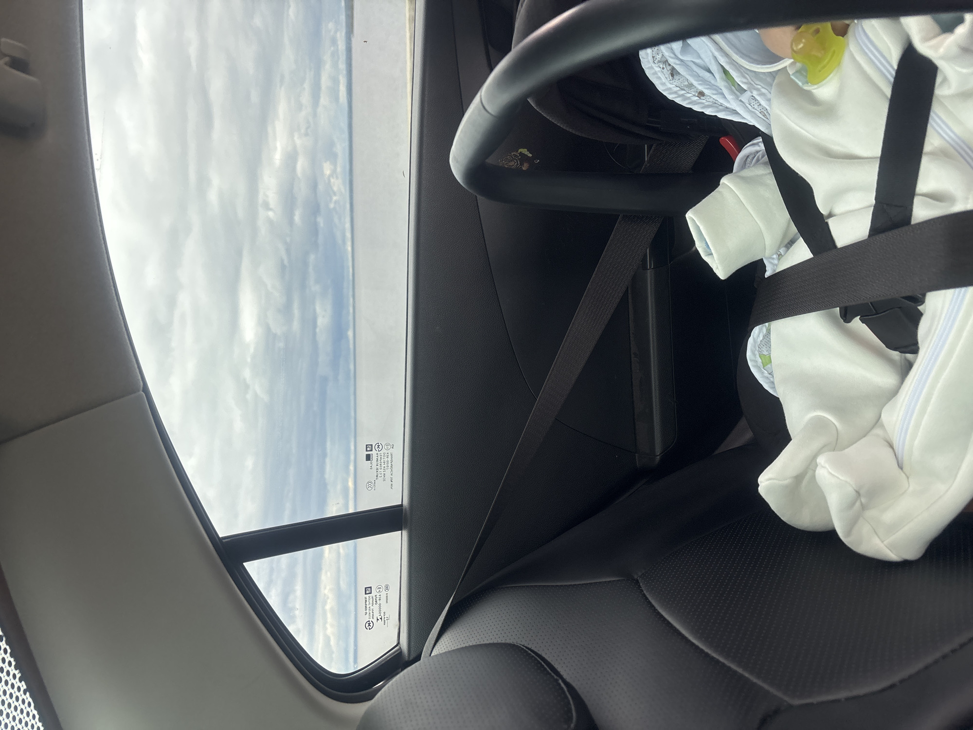 A baby in a car seat is secured with a harness in the back of a car. Outside the window is a view of the sky. The location is unknown and cannot be determined from this image.
