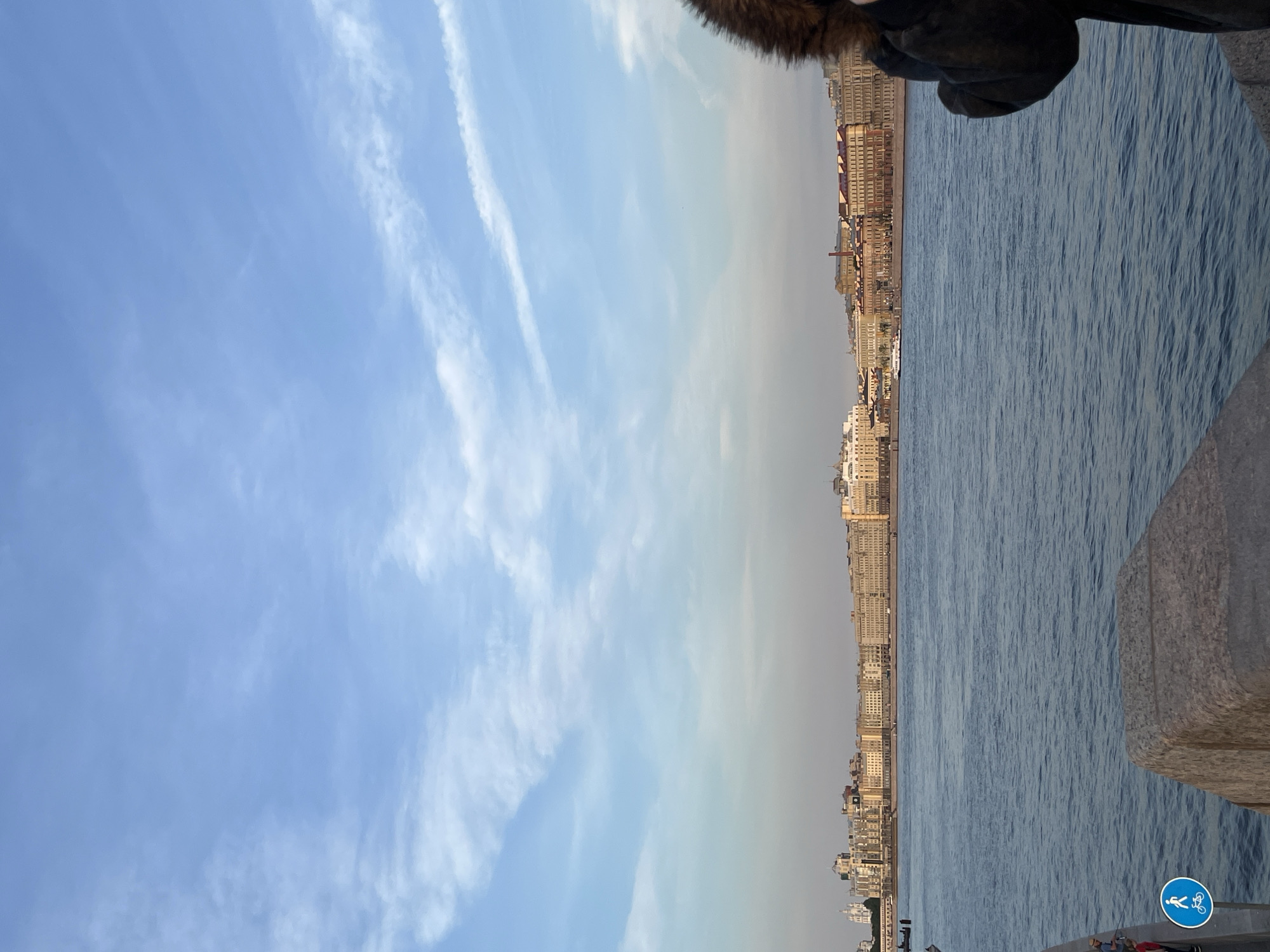 This image shows a wide shot of the Neva River in Saint Petersburg, Russia, with the historic cityscape of the city lining the opposite bank. The sky is a clear blue with wispy white clouds, suggesting it might be late afternoon or early evening. The river is a deep blue and appears calm.
On the far bank, a long line of neoclassical buildings stretches across the horizon, their facades painted in muted, warm tones. These buildings appear to be commercial or residential structures, characteristic of St. Petersburg's architectural style. There's a noticeable density of windows and architectural detailing along the facades. A small white boat is visible on the river, heading towards the left of the frame.
In the foreground, on the right side of the image, a person is partially visible, wearing a dark, fur-lined coat. Their back is mostly to the camera, and they appear to be holding something up to their face, possibly a phone or camera, looking out at the view. On the left side of the foreground, a stone embankment is visible, with two people standing near the edge. A blue circular sign with white icons of a walking person and a bicycle is mounted on a pole near them, indicating a pedestrian and cyclist path. The stone embankment itself is rough and weathered.