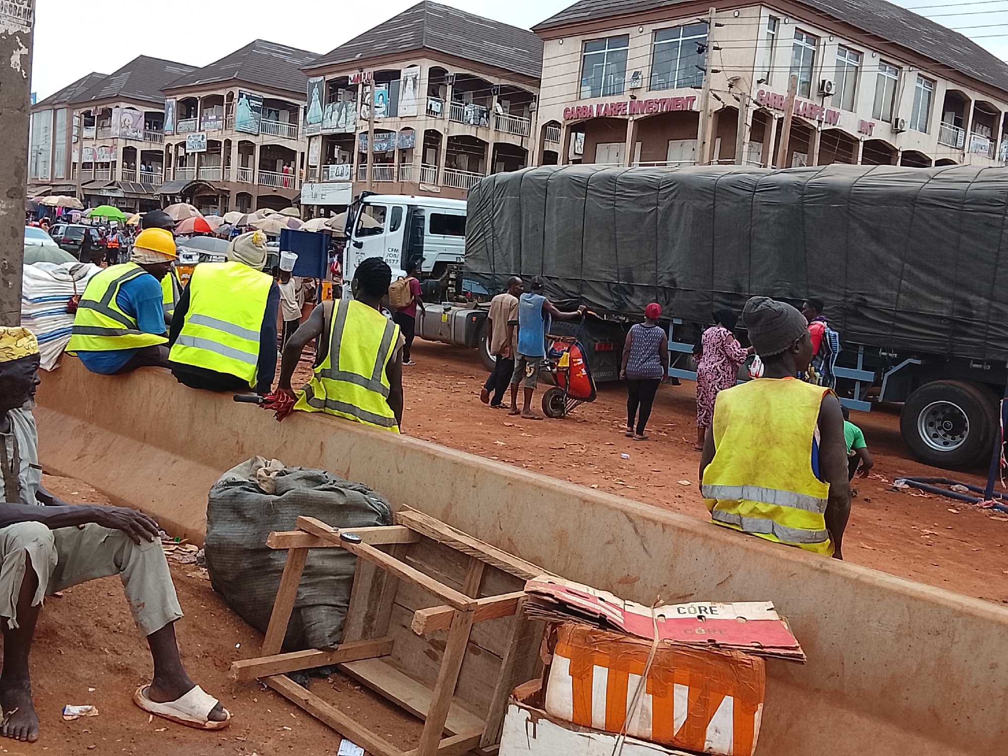 The image depicts a bustling street scene in Katambe, Nigeria. Several buildings line the background, some adorned with signage indicating businesses such as "GARBA KARFE INVESTMENT". A large truck covered with a tarpaulin dominates the right side of the frame. Numerous people are scattered throughout the scene, some wearing yellow safety vests. Several figures sit on a concrete barrier in the foreground, while others stand or walk around the truck. Debris, including a broken wooden frame and cardboard boxes, litter the ground near the barrier. The road is unpaved and appears to be a reddish-brown soil.