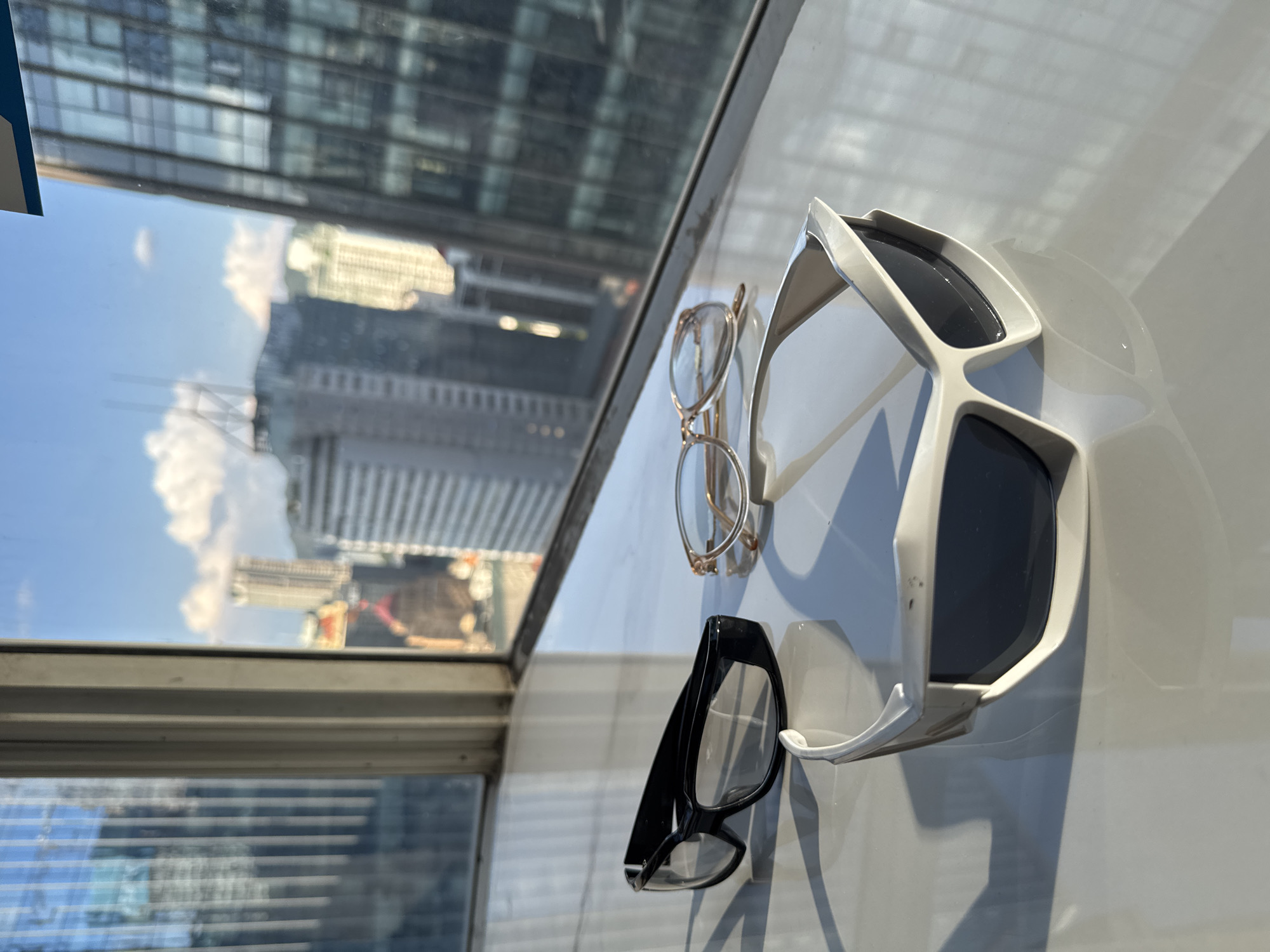 Three pairs of eyeglasses are positioned on a white reflective surface near a window. The eyeglasses are of varying styles and colors, including a black pair with clear lenses, a gold-rimmed pair with tinted lenses, and a white pair with dark lenses. The window overlooks a cityscape with tall buildings under a partly cloudy sky. The scene is illuminated by natural sunlight, casting shadows from the eyeglasses onto the surface. The location is described as the city of Yuzhong District, China. There are no people visible in the image.