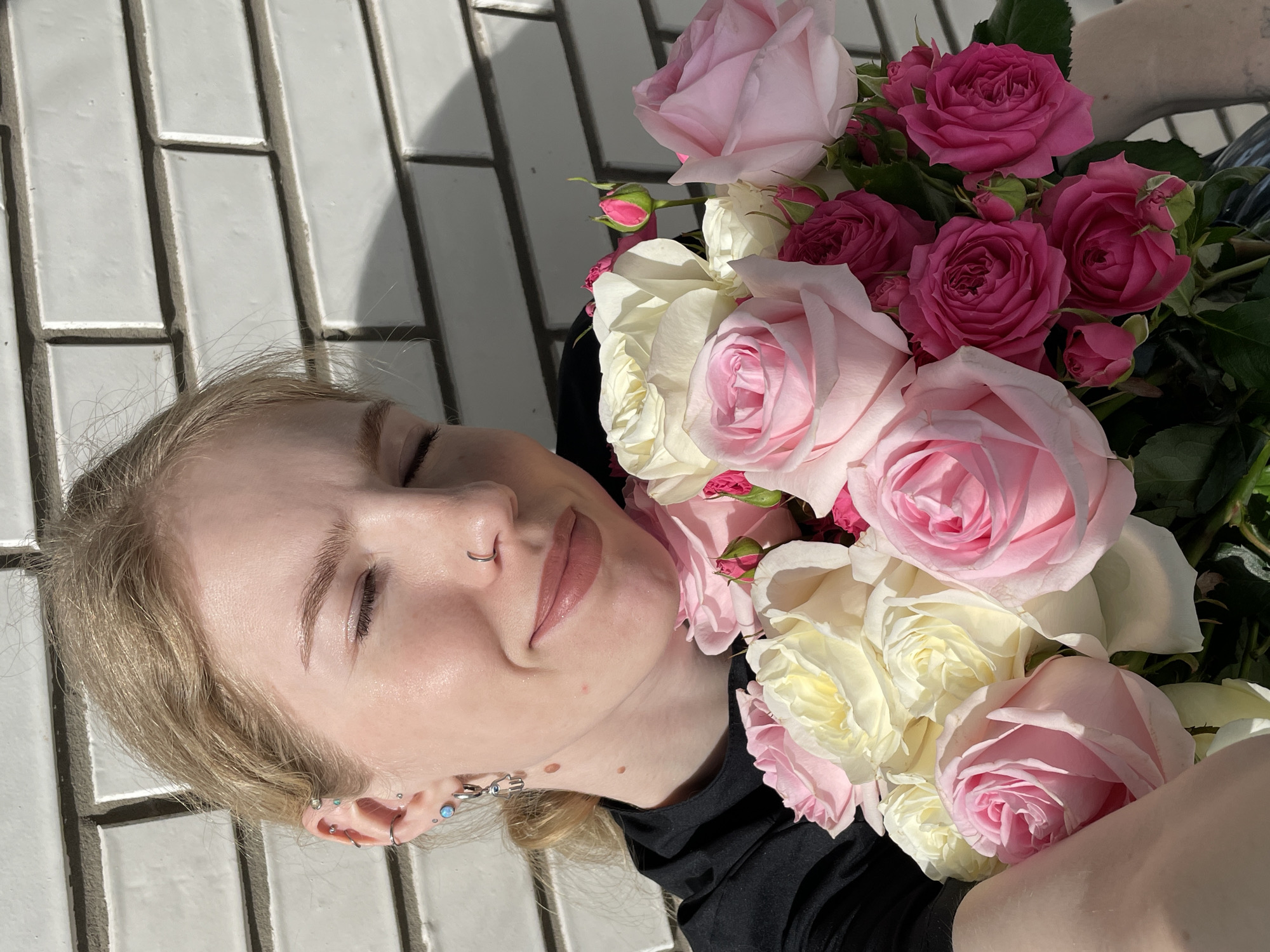 A high-angle, close-up shot features a young person lying down outdoors, framed by bright sunlight and a large bouquet of roses. The individual, appearing to be female with blonde hair, has their eyes gently closed and a subtle smile, conveying a sense of peace or contentment. They are wearing a black top.
The person's visible left ear is adorned with multiple silver earrings, including hoops and studs with iridescent blue accents, alongside a unique silver decorative earring. A silver hoop nose ring is visible on their left nostril.
A substantial bouquet of roses lies to the right, resting against the person's upper body and black clothing. The roses display a range of vibrant colors: rich fuchsia, various shades of pink (from deep to pastel), and creamy white or light yellow. Several smaller rosebuds are interspersed among the larger blooms.
The background features a light-colored, rectangular tiled surface with dark grout lines, possibly a floor or pathway. Strong, distinct shadows are cast across these tiles, indicating direct, bright sunlight. The overall atmosphere is tranquil and highlights the beauty of both the subject and the flowers. The scene is set in Izhevsk, Russia. No visible text is present in the image.