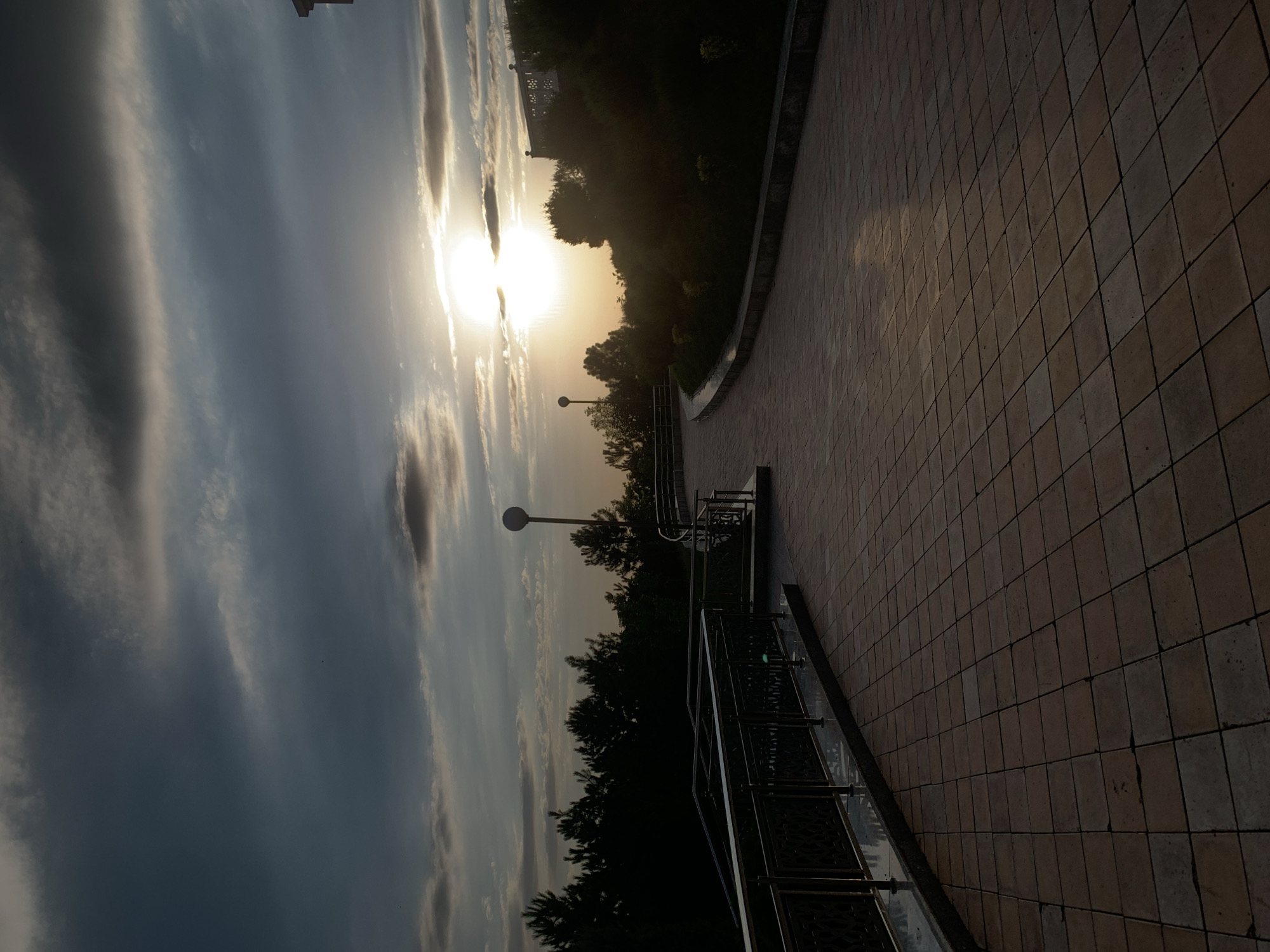 The image shows a paved walkway with square tiles, curving to the right. A metal railing runs along the left side of the walkway, with decorative metalwork visible. In the background, a dense line of dark green trees forms a silhouette against a cloudy sky. The sun is setting, partially obscured by clouds, casting a warm glow on the scene and creating reflections on the wet pavement. A single lamppost with a spherical light is visible on the left side of the walkway. On the right, a sloped area covered in dark foliage rises, with a structure at the top. The overall atmosphere is tranquil and scenic. This scene is likely located in Zaamin, Uzbekistan, a region known for its natural beauty and recreational areas.