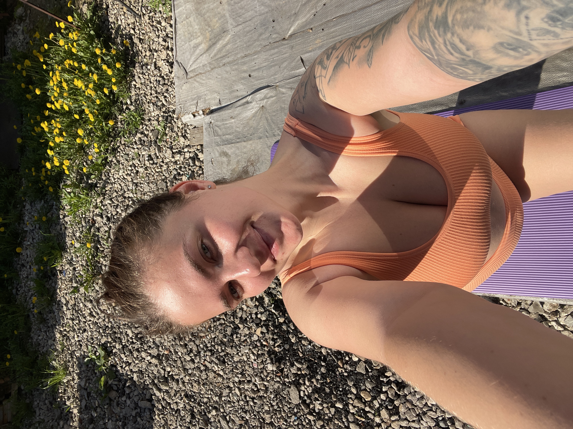 A selfie of a woman lying on a purple mat. She is wearing an orange top. Her left arm has a tattoo. The background consists of gravel, yellow flowers, and a grey tarp. The location is not discernible from the visual information provided.