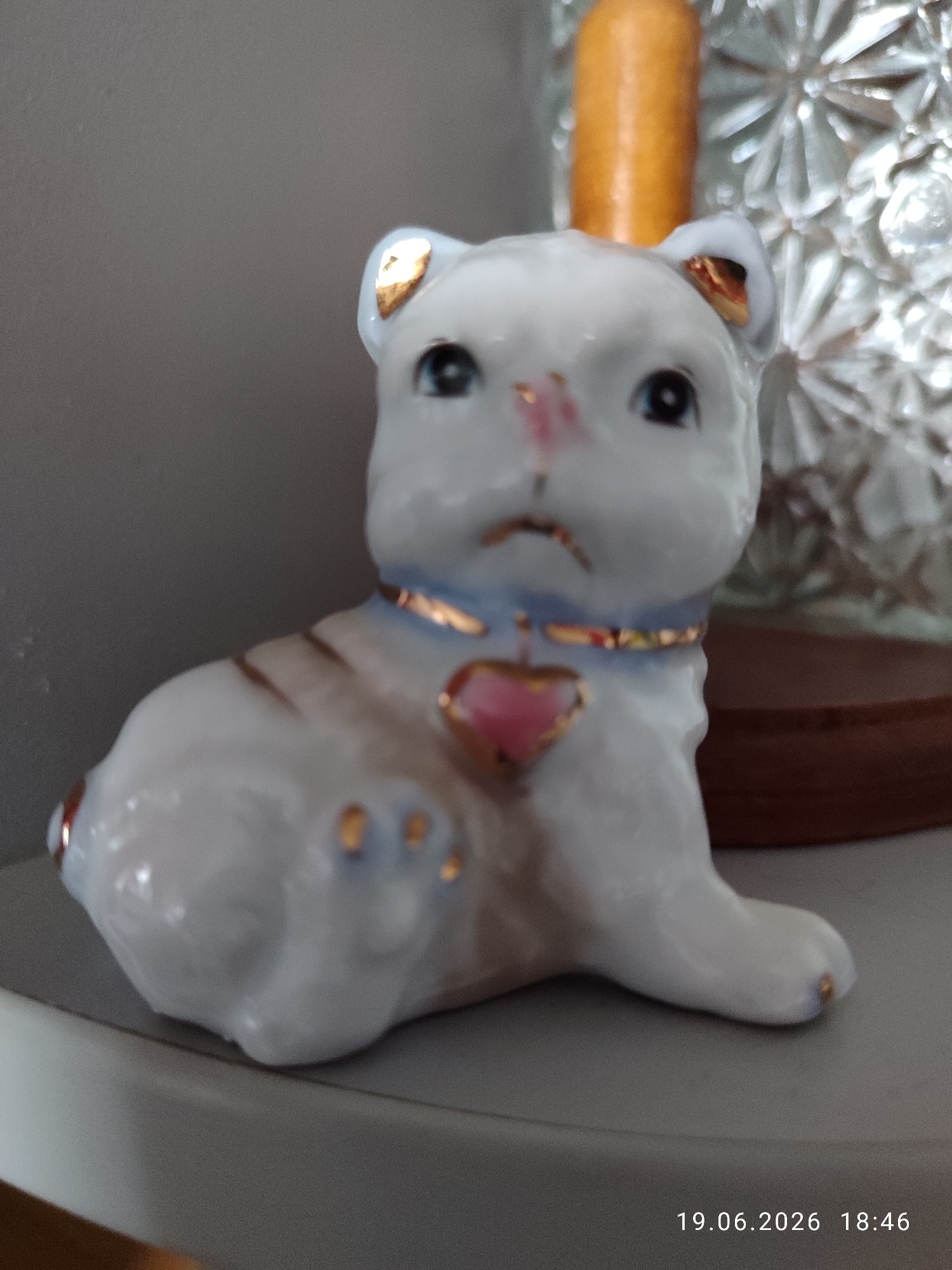 A porcelain figurine of a white cat with gold accents sits on a gray surface. The cat has blue eyes, a pink nose, and is wearing a blue collar with a pink heart pendant. Gold stripes are visible on its back and paws. In the background, a portion of a glass object with a faceted design and an orange cylindrical object are present. The scene appears to be indoors. The bottom right corner of the image shows a timestamp: "19.06.2026 18:46".