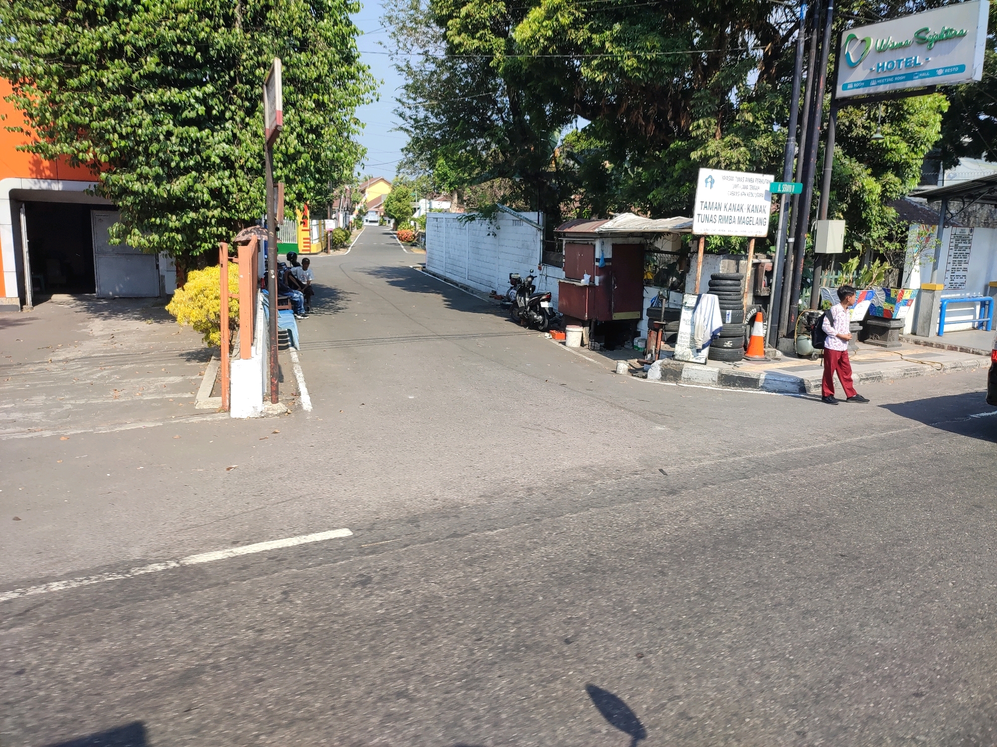 The image depicts a street scene in Magelang, Indonesia. In the foreground, a road with faded white lane markings stretches into the distance. To the right, a young person wearing red pants and a white shirt with a backpack is walking across the road. Further down the street, a small kiosk with a reddish-brown stall and a white awning is situated next to a stack of tires. A motorcycle is parked beside the stall. A white concrete wall lines the right side of the road, with trees behind it. A signpost with a green street name sign reading "JL. SERAYU II" is visible. Above the street, a large sign for "Wisma Saphira HOTEL - RESTO - ROOM - MEETING ROOM - HALL" is mounted on poles amidst lush green trees. On the left side of the road, an orange building with an open garage entrance is visible, with a person sitting on a blue chair next to it. A yellow-flowered bush and a white post with a red metal barrier are also present on the left. The overall environment appears to be a sunny day in a semi-urban area.