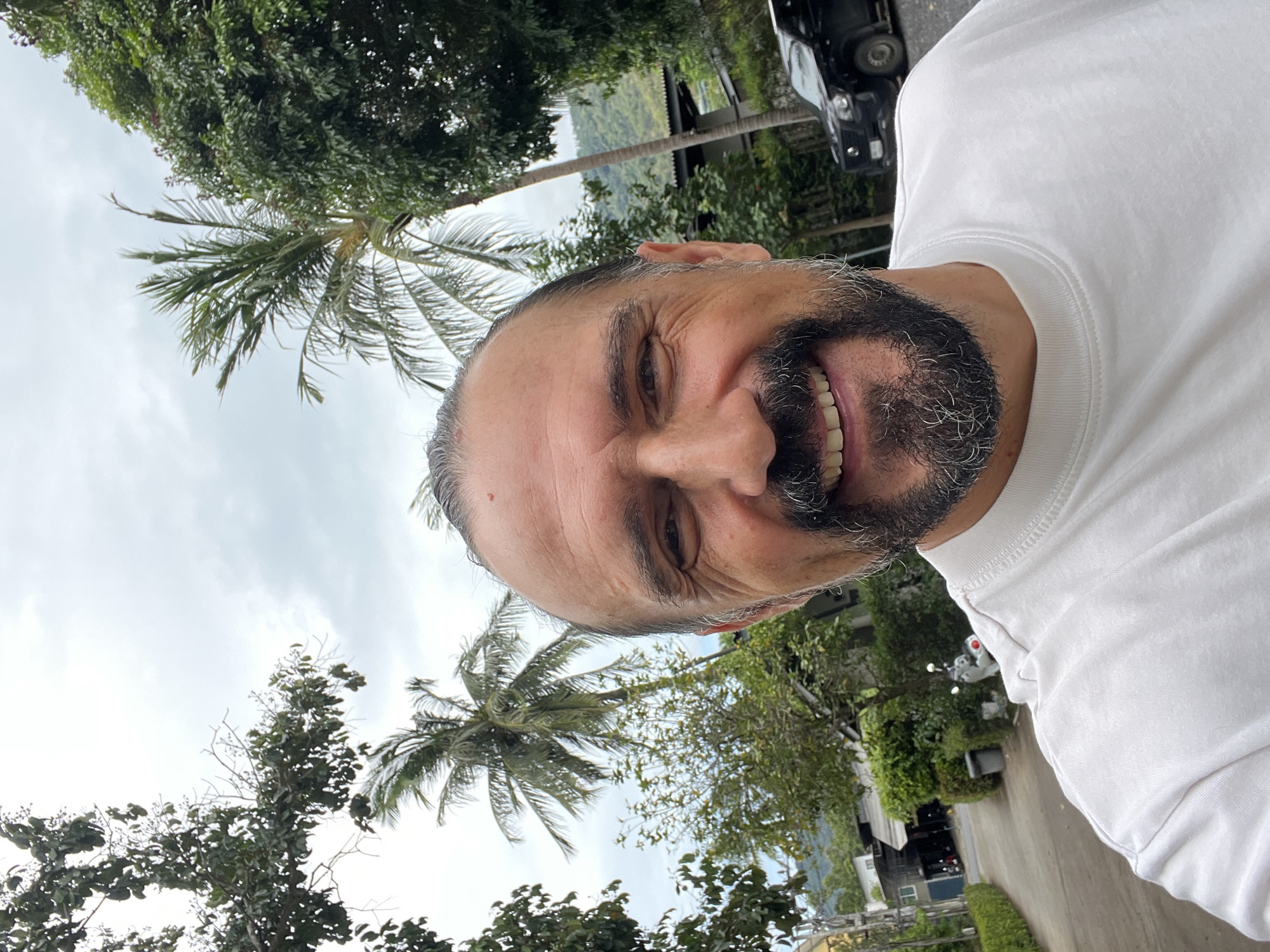 A man with a graying beard and a white t-shirt smiles at the camera. Palm trees and other greenery suggest a tropical location. A street and a black car are partially visible in the background. This scene takes place in บ้านบางรักษ์, Thailand.