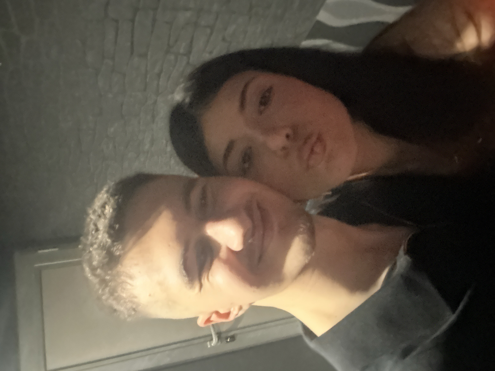 A dimly lit, close-up selfie depicts a young man and a young woman. The man is in the foreground, facing the camera with a slight smile. He has short, dark hair. The woman is positioned slightly behind him and to the side, her lips pursed in a kissing expression. Her hair is long and dark. The background consists of a patterned wall, potentially brick or wallpaper, with a window or door frame visible at the bottom left. The location is unknown and there is no information on the city of Sofia, Bulgaria.