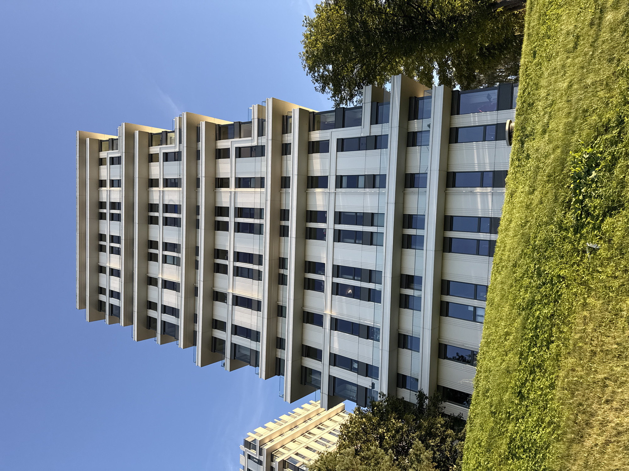 The image shows a modern, multi-story residential building with a distinctive tiered facade, located in Tallinn, Estonia. The building features a white exterior with numerous dark-framed windows and balconies with glass railings. The architecture is characterized by cantilevered sections that create a stepped appearance. To the left, another similar building is partially visible. The foreground is dominated by a lush green hillside covered in grass and vegetation. A large tree with dense foliage stands to the right of the main building. The sky is a clear, bright blue, indicating fair weather, possibly in the late afternoon given the warm sunlight illuminating the buildings. The scene depicts an urban residential area, likely a contemporary development, nestled in a natural, sloping landscape. No people are visible in the image.