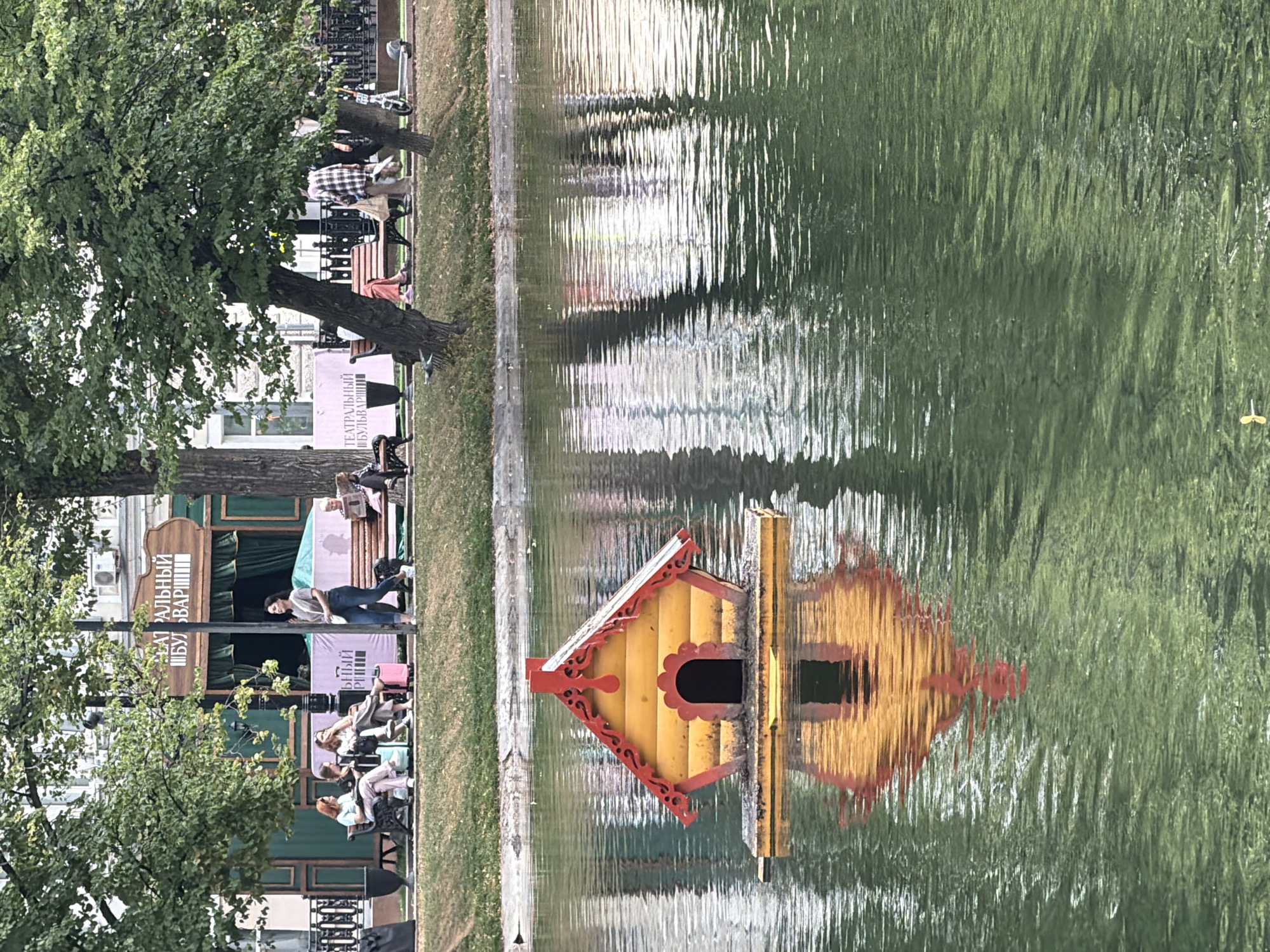 An eye-level shot captures a small, yellow and red, dollhouse-like structure floating on a body of water. The structure is square with a peaked, red roof and a small, arched doorway. Its reflection is visible on the rippling surface of the water. In the background, people are seated on benches in a park. Trees with green foliage overhang the scene, and a building with a sign featuring Russian text is visible behind the people. A lamppost stands to the right of the sign. The overall atmosphere suggests a public park in a city during daytime.