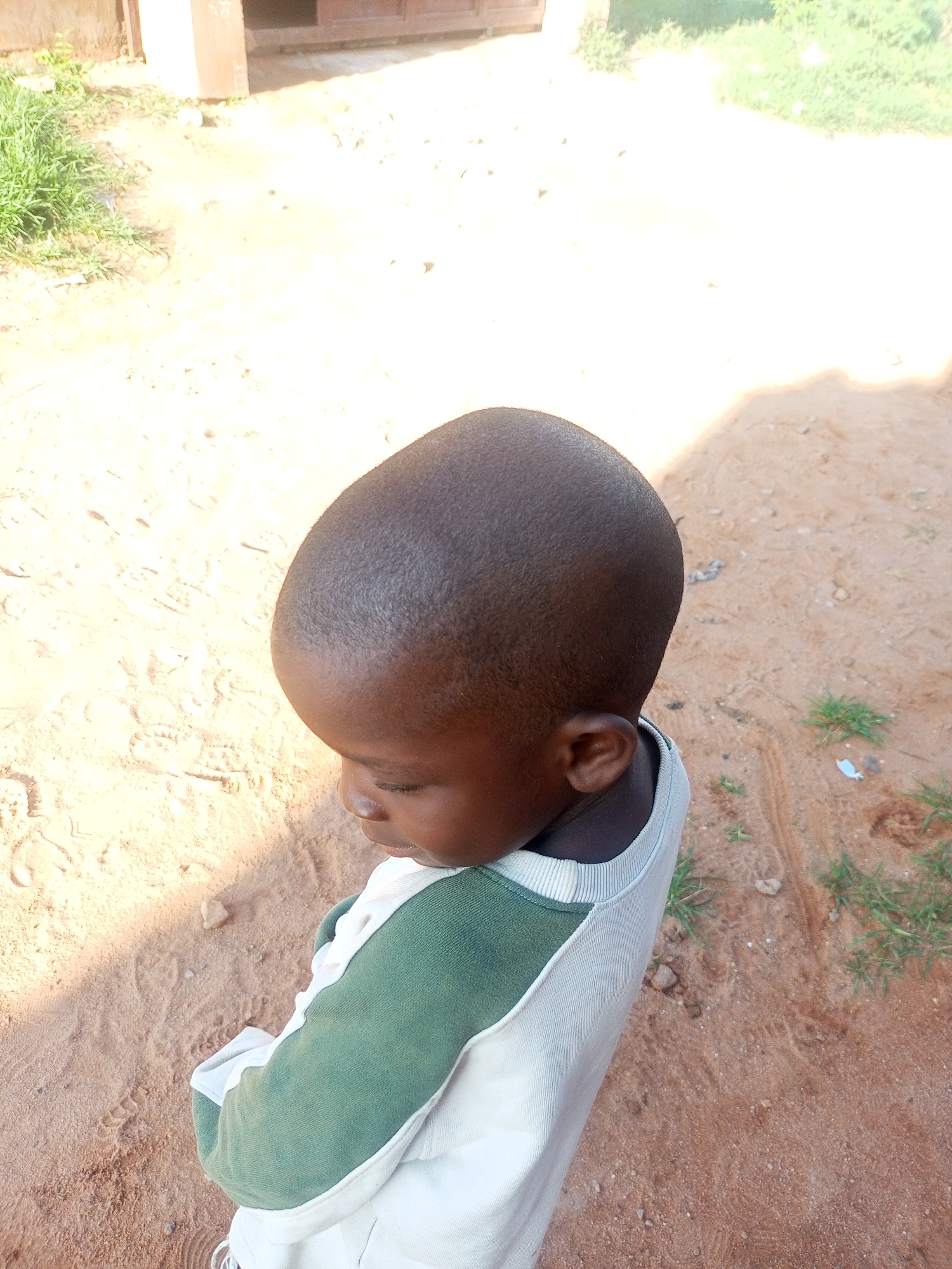 A close-up, high-angle shot shows a young Black child from the shoulders up, facing down and to the left. The child has a shaved head and is wearing a white long-sleeved shirt with green sleeves. The background is a dirt ground with some sparse grass and shadows cast by an unseen structure. The lighting is bright, suggesting it is daytime. The scene appears to be outdoors in an unpaved area.