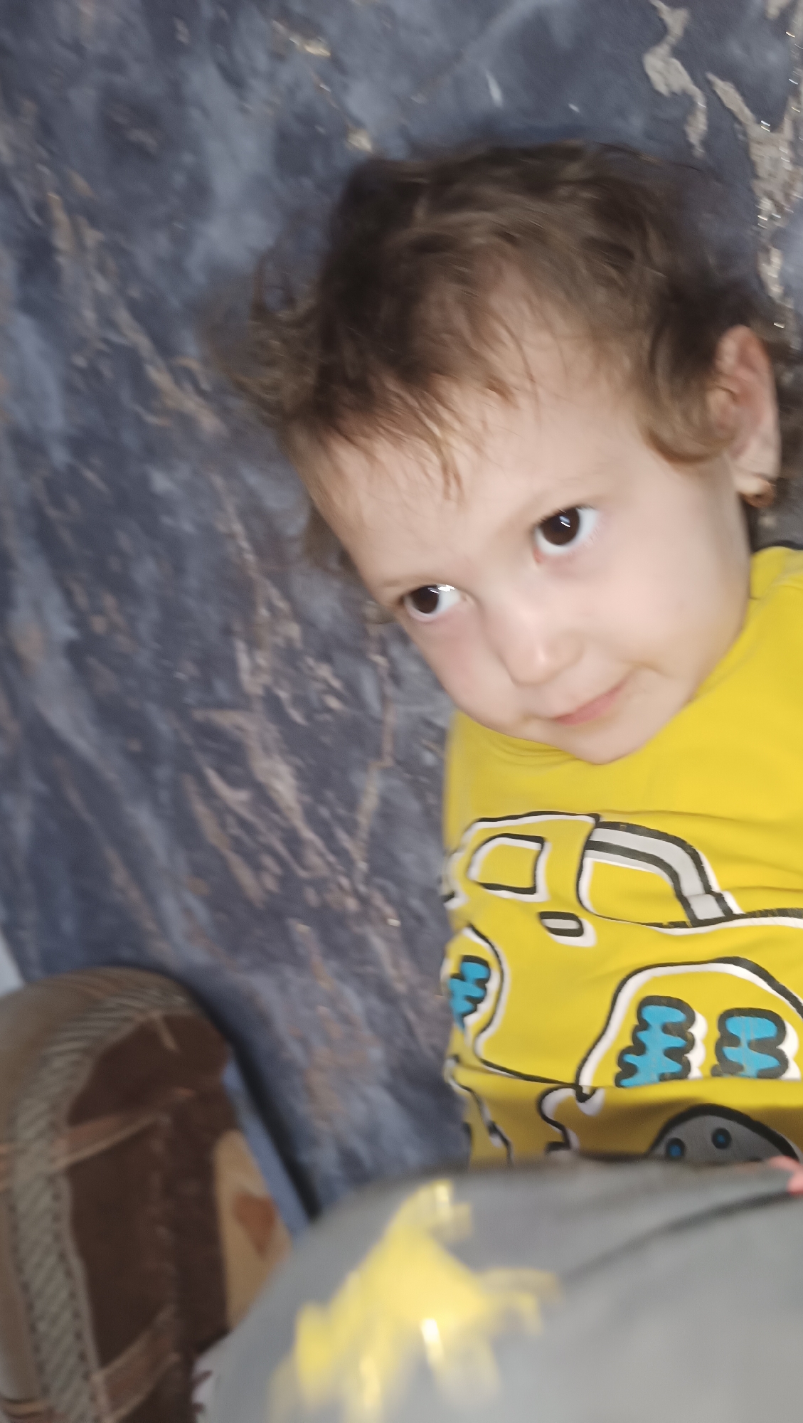 A young child with brown hair is lying on a dark, textured surface, possibly a blanket or rug. The child is wearing a yellow shirt with a graphic print of a yellow vehicle, possibly a truck or bus, with blue accents. The child's head is turned to the left, and their eyes are looking upwards. Part of the child's right foot is visible at the bottom left of the image, clad in a brown shoe with a checkered pattern. The background is a dark, mottled gray. There is no indication of the city of Karsha, Russia, in this image.