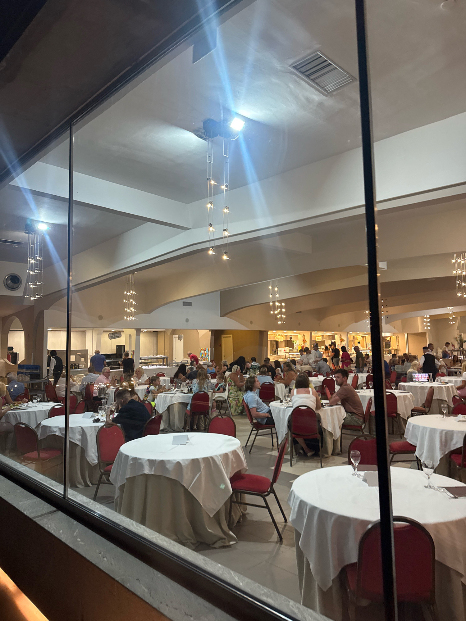 The image depicts a large indoor dining area, likely a banquet hall or restaurant, filled with people seated at round tables covered with white tablecloths. The room features high ceilings with modern chandeliers and ambient lighting. The perspective suggests the photo was taken from behind a large glass window or partition, which frames the scene and creates reflections.
In the foreground, several tables are visible, some occupied by diners. In the middle ground and background, many more tables are set up, and numerous individuals are milling about or engaged in conversation. A buffet or serving area is visible in the back, with staff and patrons around it. The architecture suggests a contemporary or renovated building.
The scene is set in Capaci, Italy. The overall atmosphere appears to be one of a social gathering, possibly a wedding reception, conference dinner, or a similar event, where a large number of people are enjoying a meal and socializing. The lighting and the reflections on the glass create a somewhat diffused and ethereal quality to the image.