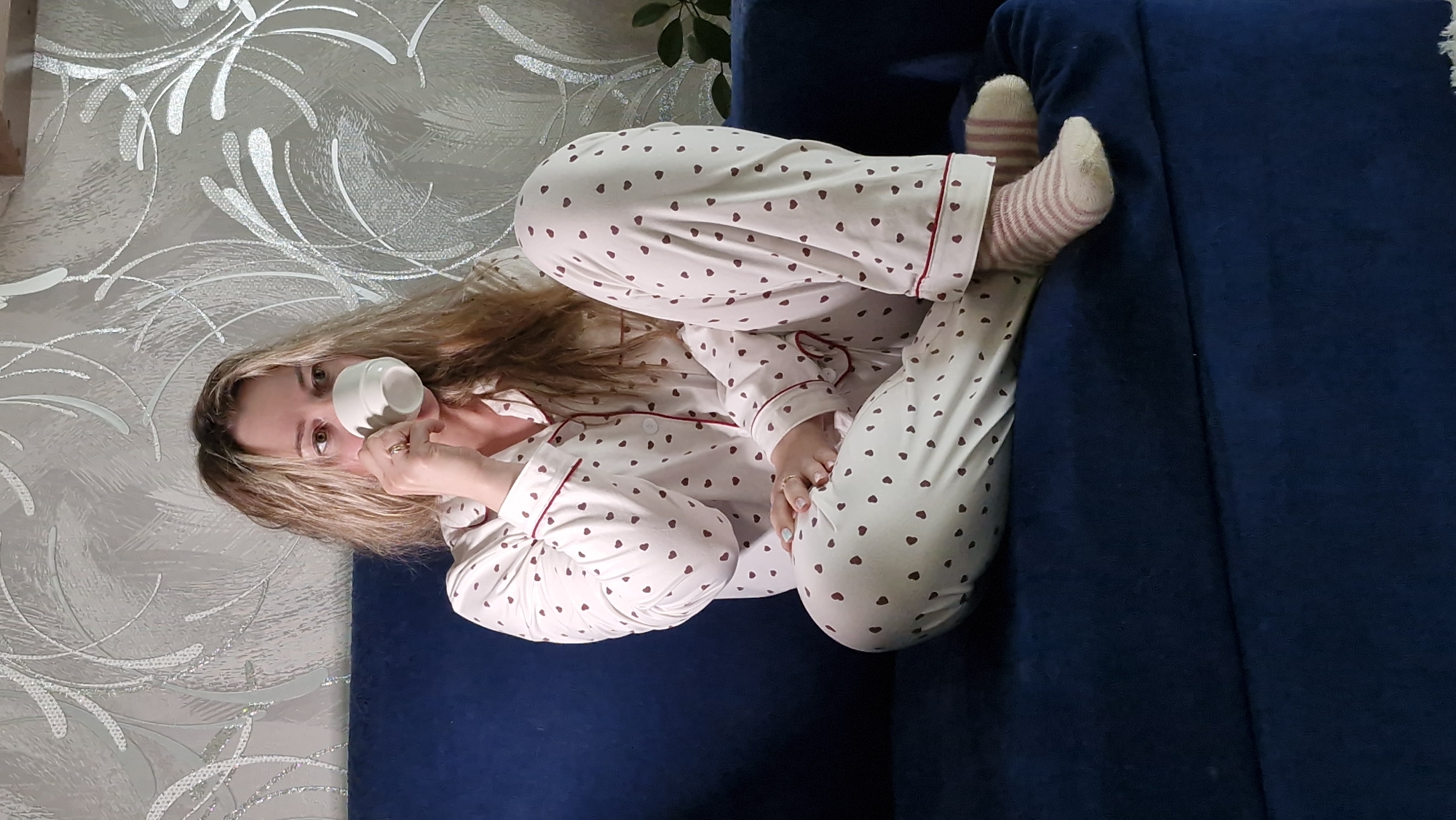 A fair-skinned woman sits cross-legged on a navy blue couch, drinking from a white mug. She is wearing white pajamas with red heart patterns and red trim, and striped socks. The background includes a wall with a silver floral pattern and a glimpse of a plant. The location is identified as Otchor, Russia, based on provided metadata.