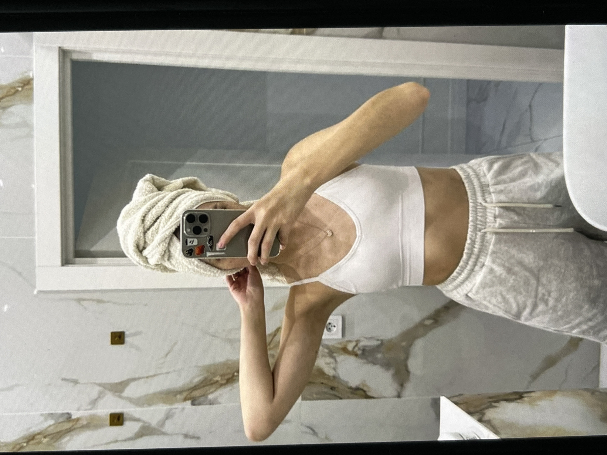 A young woman is standing in front of a mirror, taking a selfie. She has a towel wrapped around her head and is wearing a white sports bra and grey sweatpants. She is holding her phone in her right hand and appears to be adjusting her towel with her left hand. The background appears to be a bathroom with marble walls and a white door.