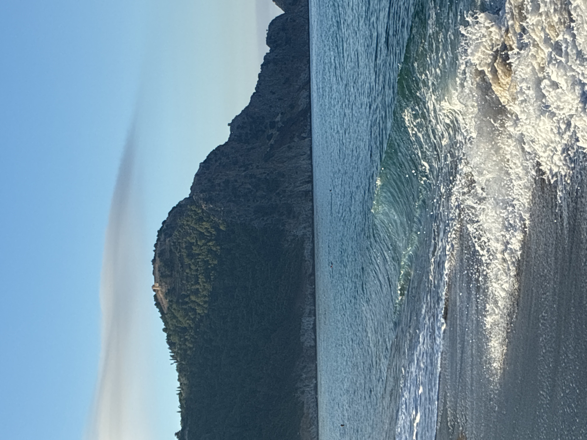 A serene seascape unfolds with a wave gently breaking on the shore in the foreground. The sea is a calm blue, leading to a lush, green mountain that dominates the background. Atop the mountain, a historical structure, likely a ruin or castle, is visible. The sky above is a clear, bright blue with a single, faint cloud stretching horizontally. The overall scene is tranquil and picturesque. The scene appears to be the Alanya coastline in Turkey, given the presence of the distinctive mountain and the visible architectural remnants often found in the region.