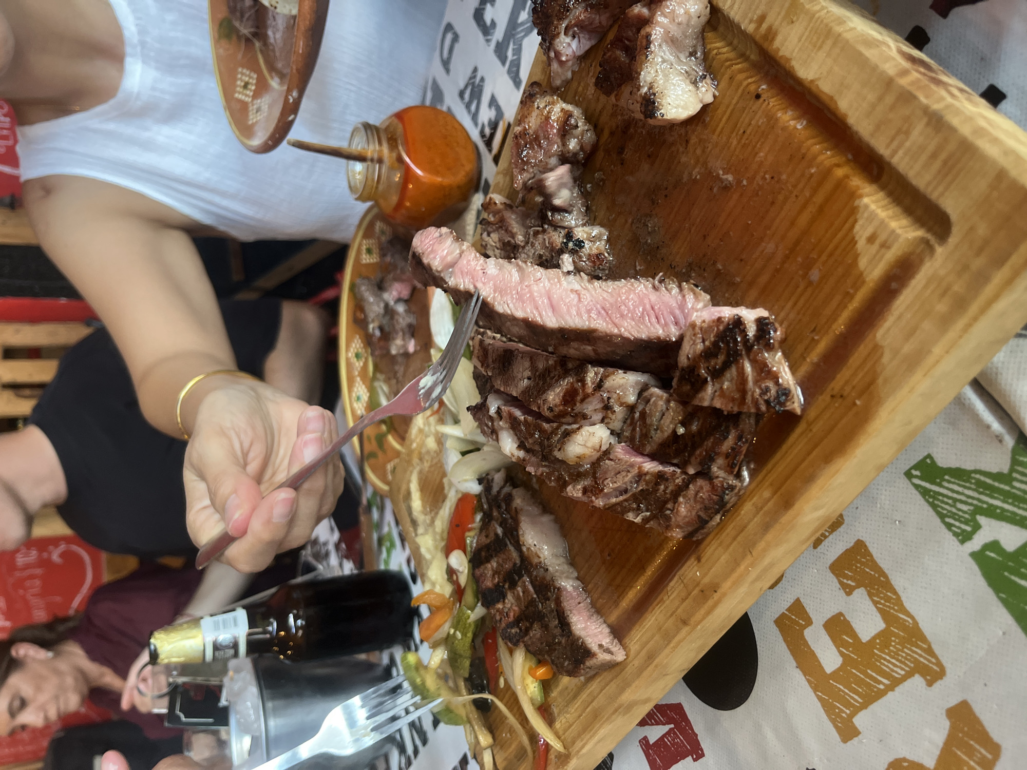 A medium-angle shot shows a person using a fork to pick up a slice of grilled steak from a wooden board laden with sliced steak and vegetables. A small glass jar filled with a reddish-orange sauce is nearby. Other items on the table include a plate, a bottle, and additional cutlery. The table is covered with a patterned tablecloth. The location may be San Francisco Totimehuacán, Mexico, though there is no explicit indication in the image.