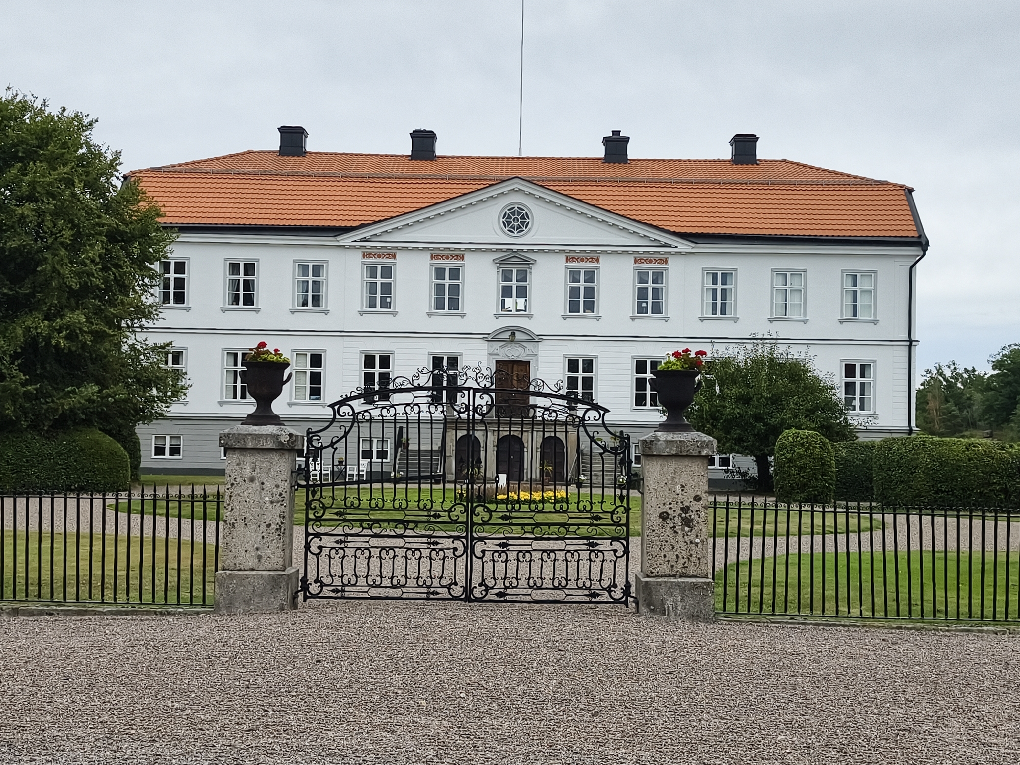 The image shows a stately white building with an orange tiled roof, likely a manor or mansion, located in Brokind, Sweden. The building features multiple windows with white frames and an elaborate pediment above the central section, which includes a decorative circular window. In front of the building is a black wrought-iron gate, flanked by stone pillars. The gate opens onto a manicured lawn with a gravel driveway in the foreground. Two large planters with red flowers are placed on top of the stone pillars. A black metal fence extends from the pillars, enclosing the property. Lush green trees and hedges are visible to the left and right of the building, and in the background. The sky is overcast. There are no people or animals present in the image.