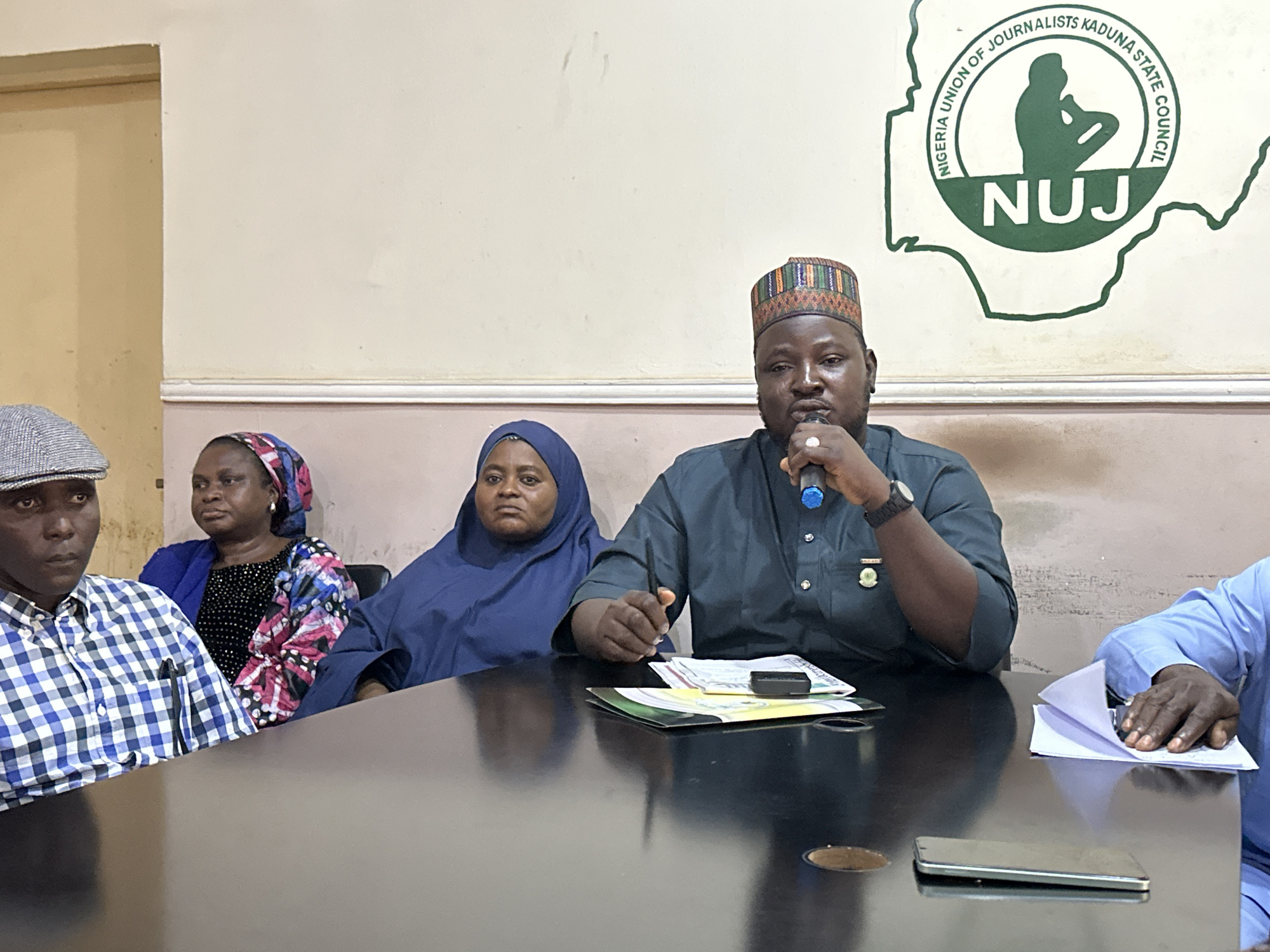 A medium shot captures a group of four individuals seated at a dark, reflective table in a room. The setting is likely an official meeting, as indicated by a prominent circular logo on the wall behind them featuring the text "NIGERIA UNION OF JOURNALISTS KADUNA STATE COUNCIL" and "NUJ." The logo is rendered in green on a pale, textured wall.
On the left, a man wearing a grey and white checkered shirt and a grey cap is partially visible, looking to his left. To his right, a woman in a blue headscarf and a colorful, patterned garment is seated, looking forward. Next to her, a woman in a blue hijab is positioned, also looking ahead. The central figure is a man wearing a dark green traditional outfit and a colorful, patterned cap. He is holding a microphone in his right hand and speaking, with a ring on his left hand. His right arm rests on the table, near a stack of papers and a small black device. On the far right, another person, wearing a light blue traditional outfit, is partially visible, with their hands resting on a stack of white papers. The table itself is cluttered with papers, a phone, and a circular cutout that reveals a darker surface below. The lighting appears to be artificial, casting a warm glow on the scene.