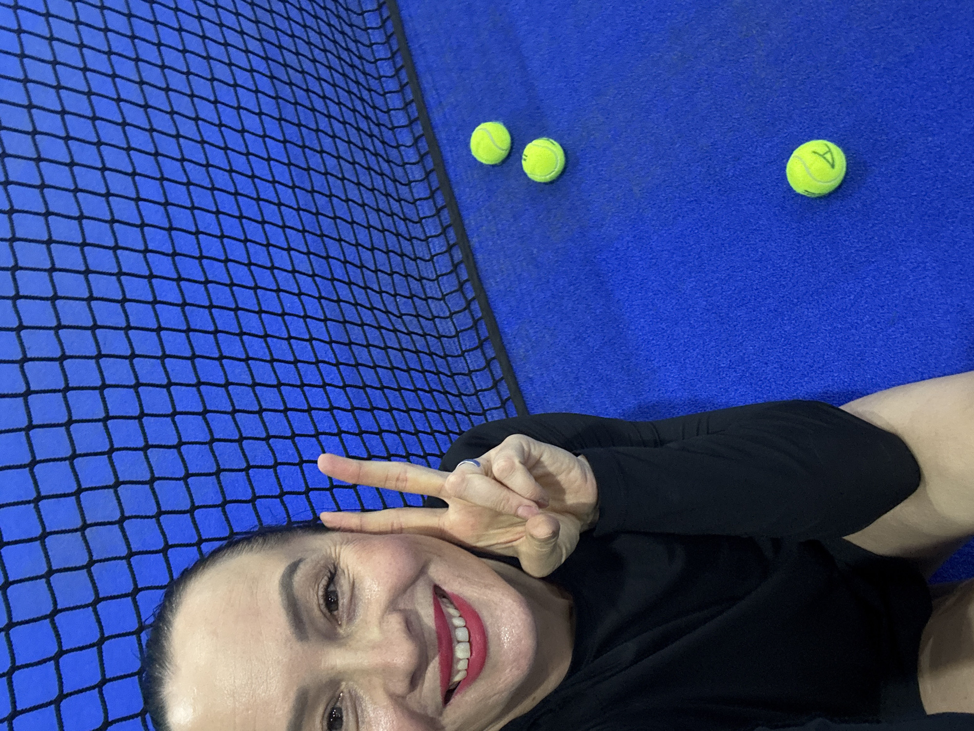 A woman is taking a selfie indoors on a blue surface next to a black net, with three tennis balls in the background. The woman is smiling and making a peace sign with her fingers. She is wearing a black long-sleeved shirt. The location could be the city of Chaikovsky, Russia.