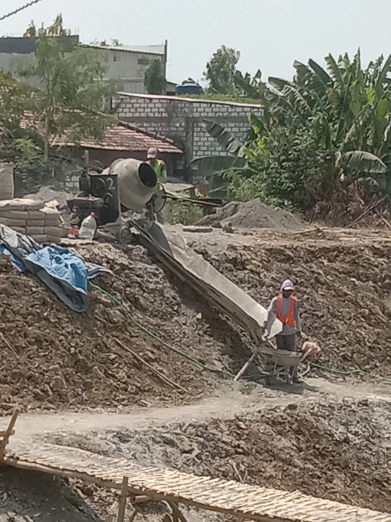 The image depicts a construction site in Made Kampung, Indonesia. A cement mixer is visible in the background, next to stacks of cement bags. A worker, wearing a high-visibility vest and a cap, is operating a wheelbarrow on a dirt embankment. Another person in a similar vest is near the cement mixer. The environment is characterized by piles of dirt, construction materials, and vegetation, including banana trees. A wooden plank walkway is present in the foreground. The scene suggests ongoing construction activity.