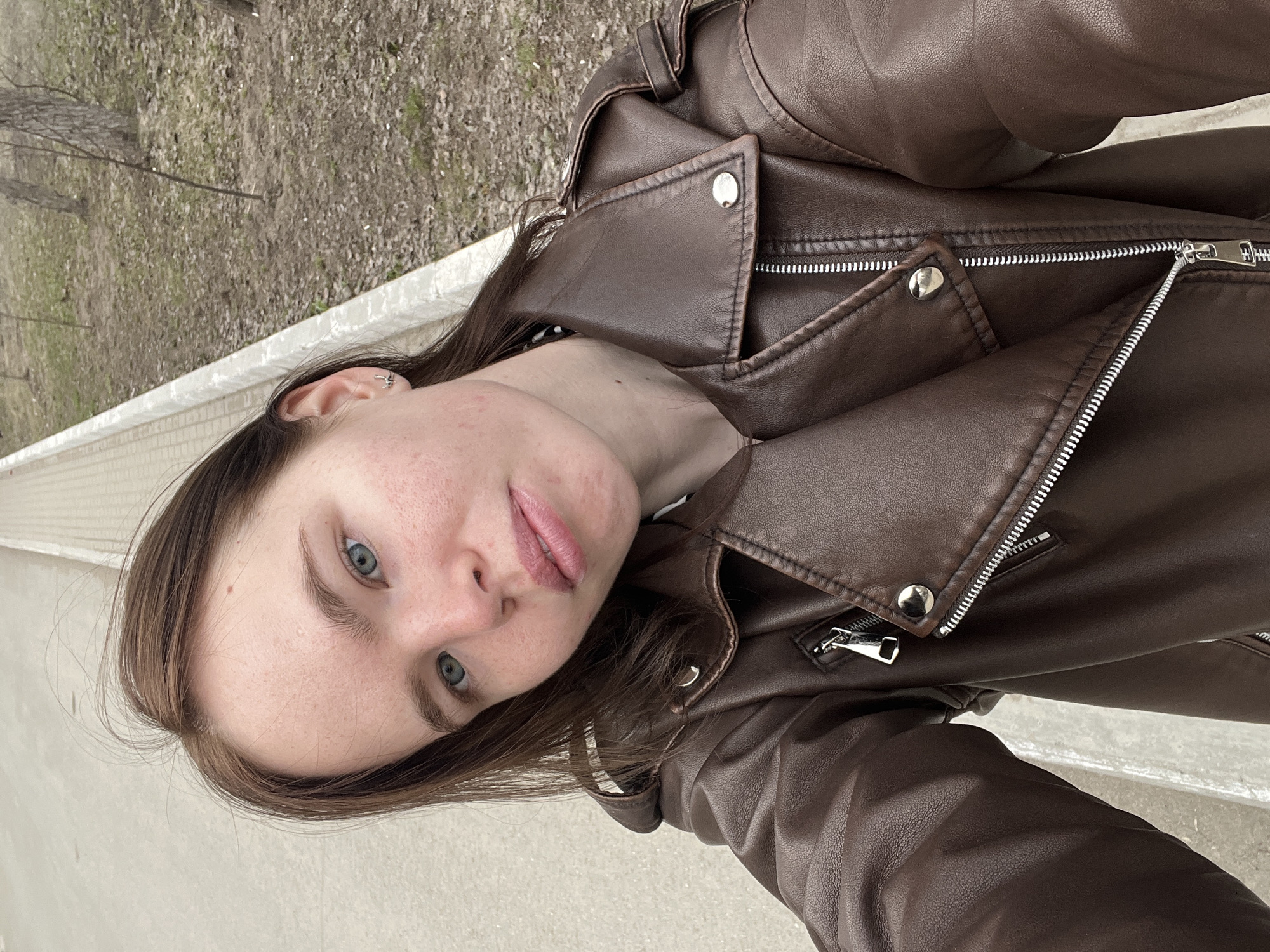 A person with light skin, brown hair, and blue eyes is taking a selfie outdoors. They are wearing a brown leather jacket. The background shows a concrete structure, possibly a retaining wall, and a bare tree branch suggesting a park or outdoor area in a colder climate.
It is not possible to provide the specific location of the city of Chaikovsky, Russia based on the provided image.