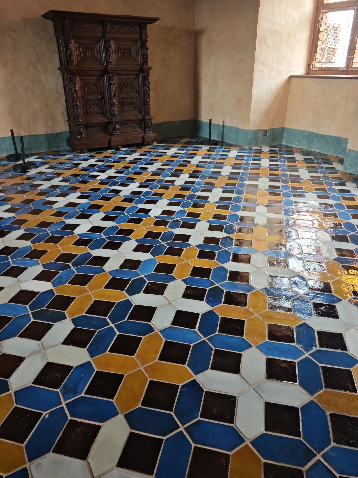 The image depicts the interior of a room with a highly decorative tiled floor. The tiles are geometric in pattern, featuring a repeating arrangement of octagons and squares in colors of blue, yellow, white, and dark brown. A tall, intricately carved wooden cabinet stands against a textured, pale peach-colored wall on the left side of the room. The wall has a lower section painted in a dark teal color. A window with a diamond-patterned grid is visible in the upper right corner, letting in natural light that reflects off the polished floor. The overall impression is of a historical or museum setting. The location is the Bauska Castle complex in Bauska, Latvia. There are no people visible in the image.