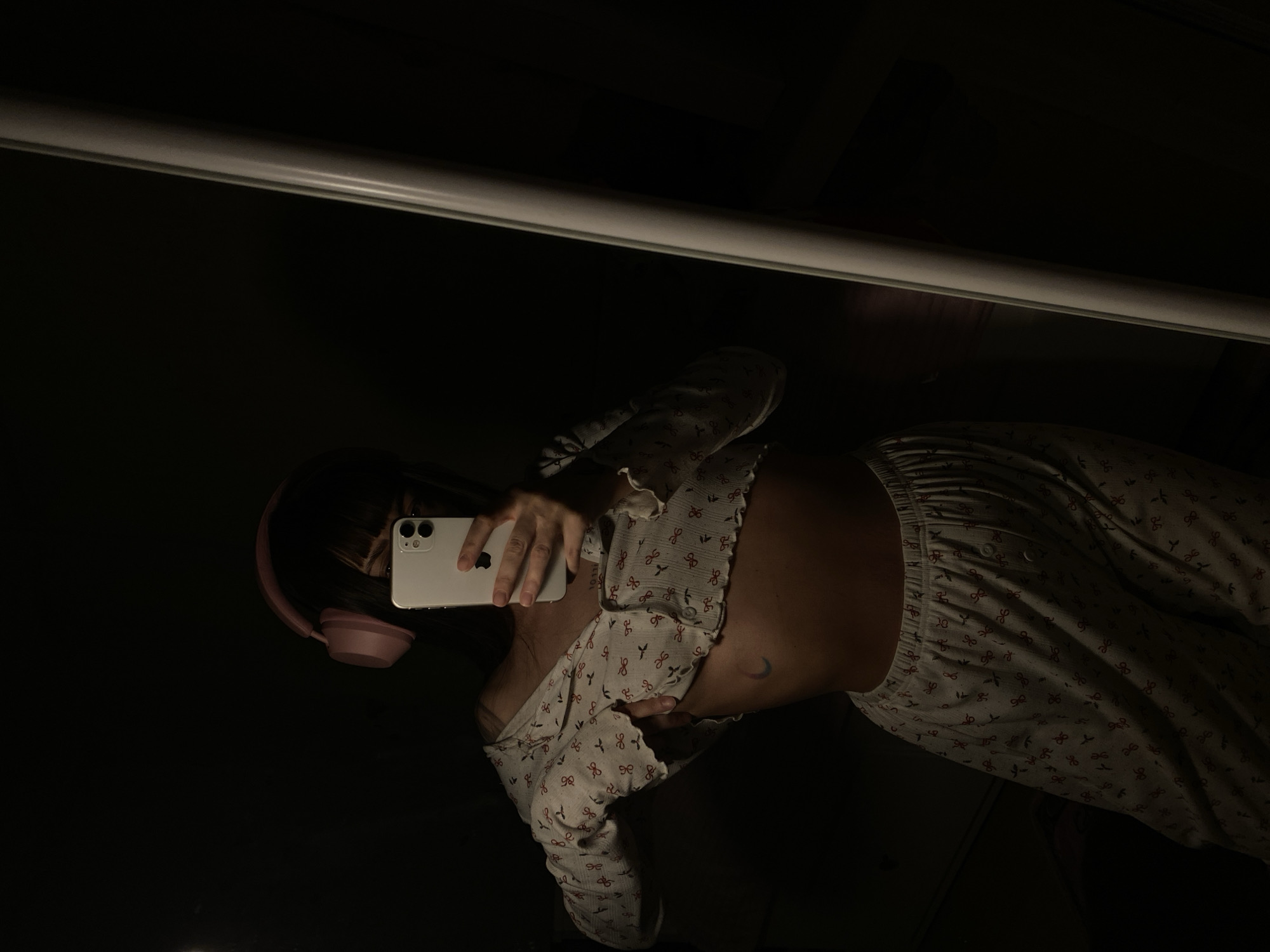 A dimly lit selfie is being taken in a mirror. A person, wearing headphones, pajamas with a red bow pattern, and a top with similar patterns is seen holding a white smartphone. The background is dark, with a blurry white light fixture visible at the top of the frame. It is not possible to ascertain the location as Votkinsk, Russia based on the visual information.