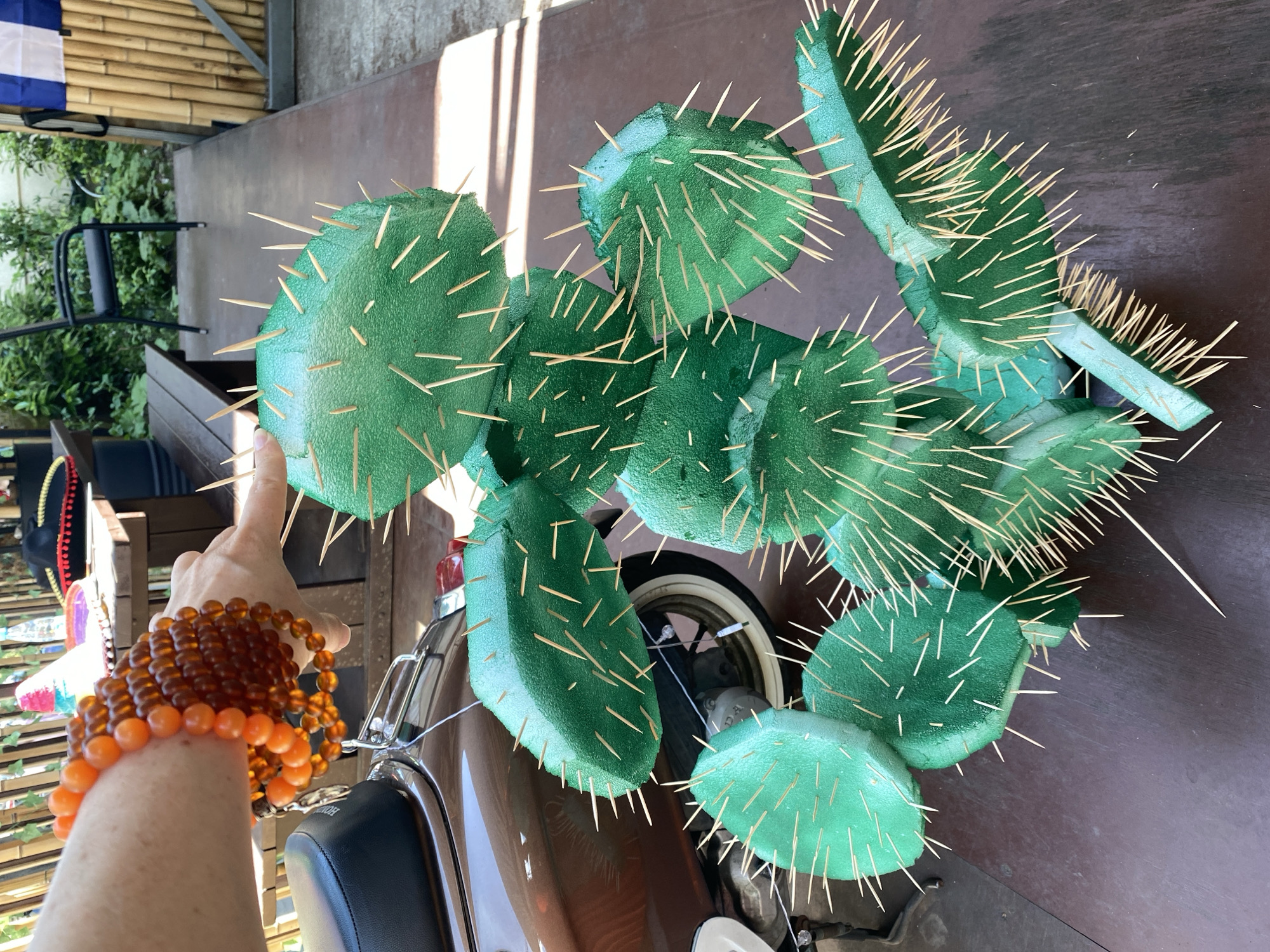 The image depicts an outdoor scene in Sochi, Russia, featuring a human arm interacting with a large, decorative green cactus.
In the upper left quadrant, a fair-skinned human forearm and hand are visible. The forearm wears multiple strands of spherical beaded bracelets; two strands are composed of opaque orange beads, and several other strands consist of translucent amber-colored beads. The index finger of the hand is extended, pointing directly towards the top-most segment of a prominent green decorative cactus.
The central subject is a multi-segmented, artificial cactus, fabricated from a light green foam-like material. It is designed to resemble a prickly pear cactus (Opuntia species). Numerous thin, light-colored wooden spikes, resembling toothpicks, are embedded into the green foam segments, simulating thorns. The cactus stands upright on a dark brown wooden deck or planked floor.
In the mid-left, partially obscured by the cactus and the human arm, is the rear section of a brown scooter or small motorcycle. Visible components include a black seat, chrome handlebars/frame, and a partial view of a white-walled wheel and tire.
The background, particularly in the upper center and right, shows elements of an outdoor patio or garden area. A wooden table with a light-colored, possibly straw, sombrero and a clear bottle is visible. Further back, green foliage and a black metal chair indicate a natural outdoor setting. To the far right, a vertical bamboo fence or screen is present, with a fragment of blue and white striped fabric (possibly a flag or banner) hanging from it.
The entire scene is illuminated by natural light, casting distinct shadows on the dark wooden floor, suggesting an outdoor environment on a sunny day.