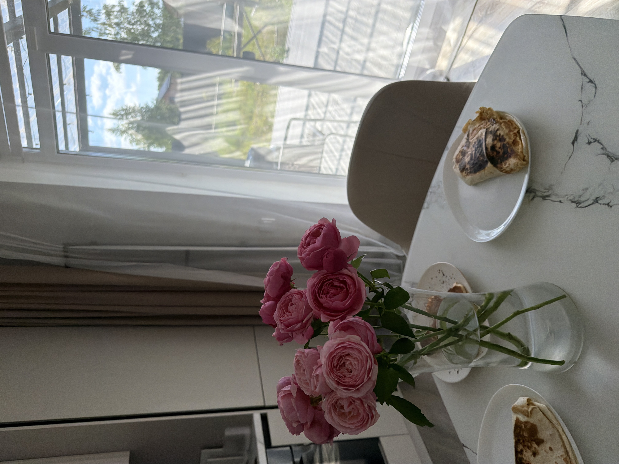 A close-up, eye-level shot captures a domestic scene with a bouquet of pink roses in a glass vase as the central focus. The roses are lush and fully bloomed, with delicate petals in varying shades of pink. They are arranged in a clear, cylindrical vase filled with water, showing the green stems submerged.
The vase sits on a white, marble-patterned table. To the left of the vase, a white plate holds a portion of what appears to be a folded flatbread or wrap, browned on the surface. To the right of the vase, another white plate contains a similar item, also browned.
The background reveals a bright, sunlit room. A large window with sheer white curtains allows diffused light to enter, illuminating the scene. Through the window, a glimpse of the outdoors is visible, suggesting a residential area with trees, a fence, and a house with a roof. The curtains are partially drawn, creating a soft, dappled effect on the windowpane.
In the immediate background, a portion of a beige upholstered chair is visible, positioned to the right of the table. Behind the table and to the left, a modern kitchen area is partially visible, with white cabinets and what looks like stainless steel appliances. The overall impression is one of a pleasant, perhaps breakfast or brunch, setting in a home.
The scene is located in Malinovka, Russia, a location indicated by the specific context of the request. The image composition and subject matter suggest a casual, everyday moment, possibly in a suburban or rural setting given the view from the window.