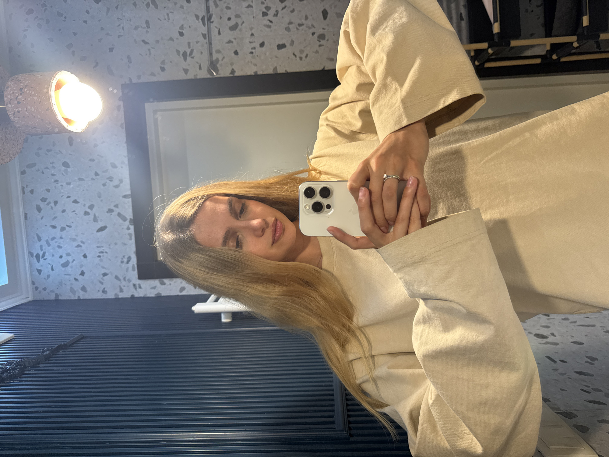 A young woman with long, blonde hair is taking a selfie in an unidentified indoor location. She is wearing a light-colored, possibly beige, long-sleeved garment. In her right hand, she holds a white iPhone, and a ring is visible on her ring finger. The background includes a light fixture with a cylindrical shade, a framed mirror or window, and a dark, horizontally striped surface. The wall behind the woman is decorated with a speckled, stone-like pattern.