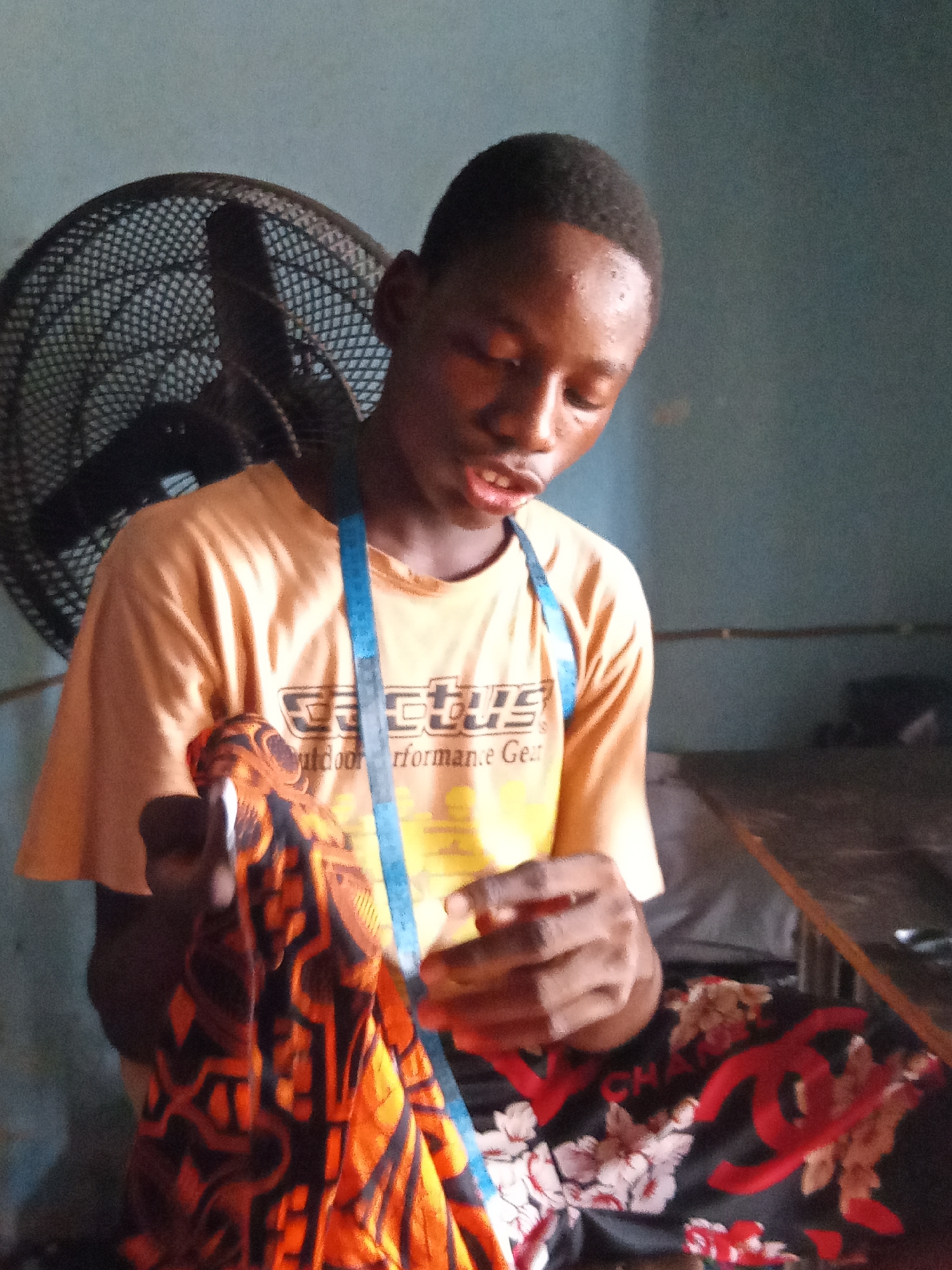 A young man stands indoors, holding a piece of patterned orange and black fabric. He wears a light orange t-shirt with text on the front and a blue measuring tape around his neck. Behind him is a black fan. He is wearing black pants with red and white designs. The scene is situated near Nsugpe, Nigeria.