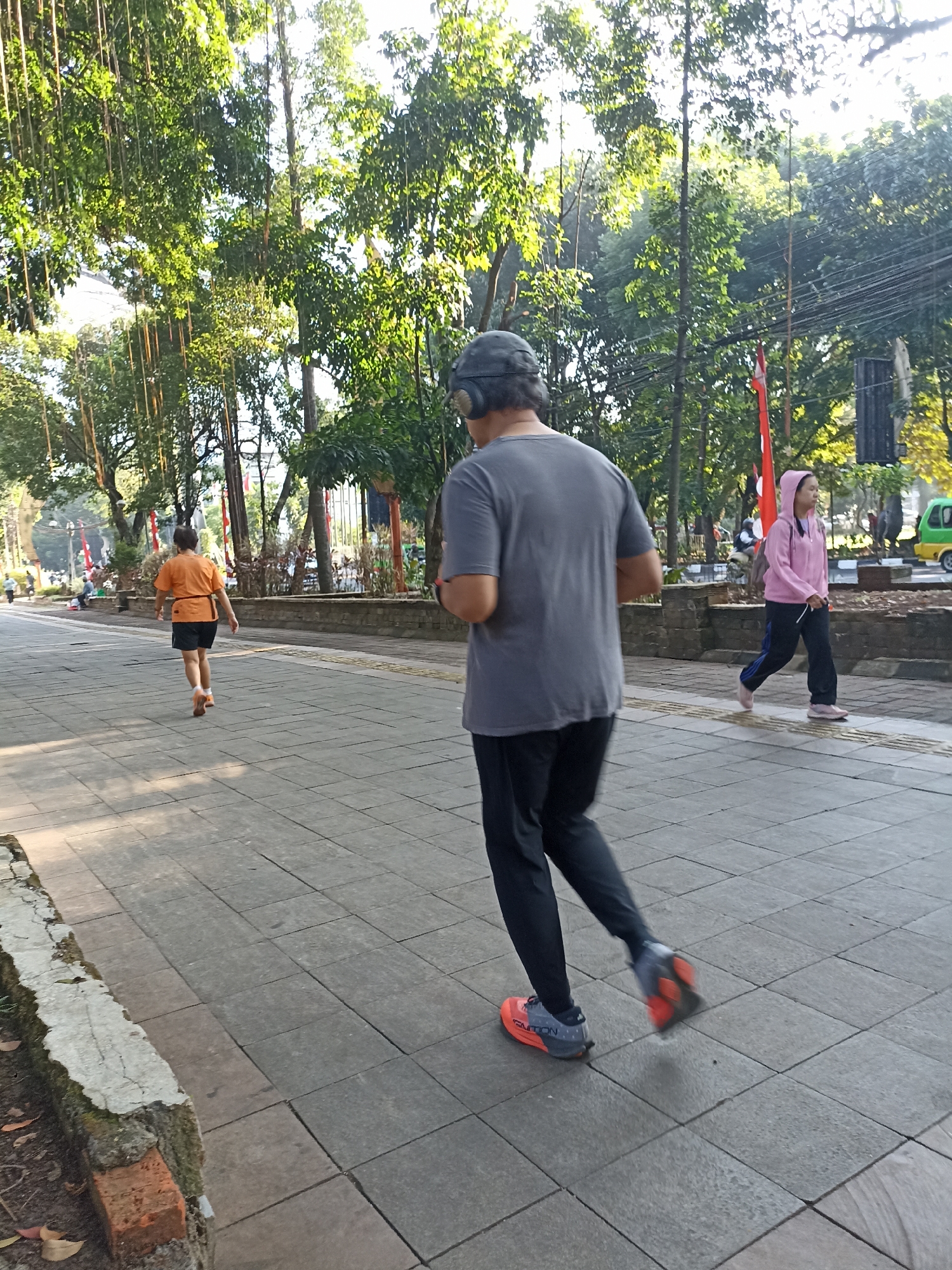 The image depicts a paved walkway in a park-like setting, likely in Bogor, Indonesia, given the presence of lush, tropical trees with hanging roots. Sunlight filters through the canopy, casting dappled light on the pathway. Three individuals are visible, all engaged in physical activity. In the foreground, a person wearing a grey t-shirt, black pants, and headphones is jogging away from the camera. To the left, a person in an orange t-shirt and black shorts is also jogging ahead. Further in the background, on the right, a person in a pink hooded jacket and black pants is walking. The walkway is made of rectangular grey tiles, with a brick and concrete border along the left edge. A stone wall runs along the right side of the walkway, behind which are more trees and some red flags. Electrical wires are visible strung between poles in the background, suggesting an urban or semi-urban park environment.