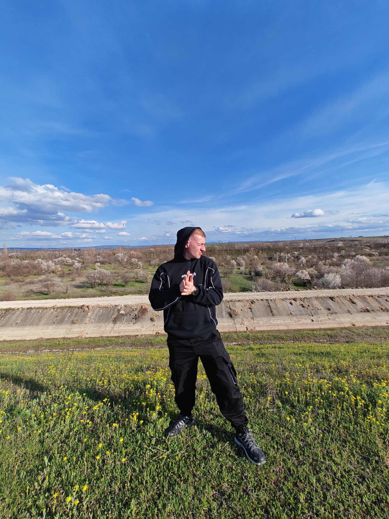 A medium shot captures a fair-skinned man dressed in black against a landscape under a partly cloudy sky. The man wears a hooded sweatshirt with white accents and black cargo pants, paired with grey and black athletic shoes. He stands in a grassy field speckled with yellow wildflowers, positioned in front of a concrete irrigation canal. Beyond the canal, a rural landscape stretches to the horizon, dotted with sparse vegetation and flowering trees. The sky is blue with scattered white clouds. This scene is in Shumen, Bulgaria.