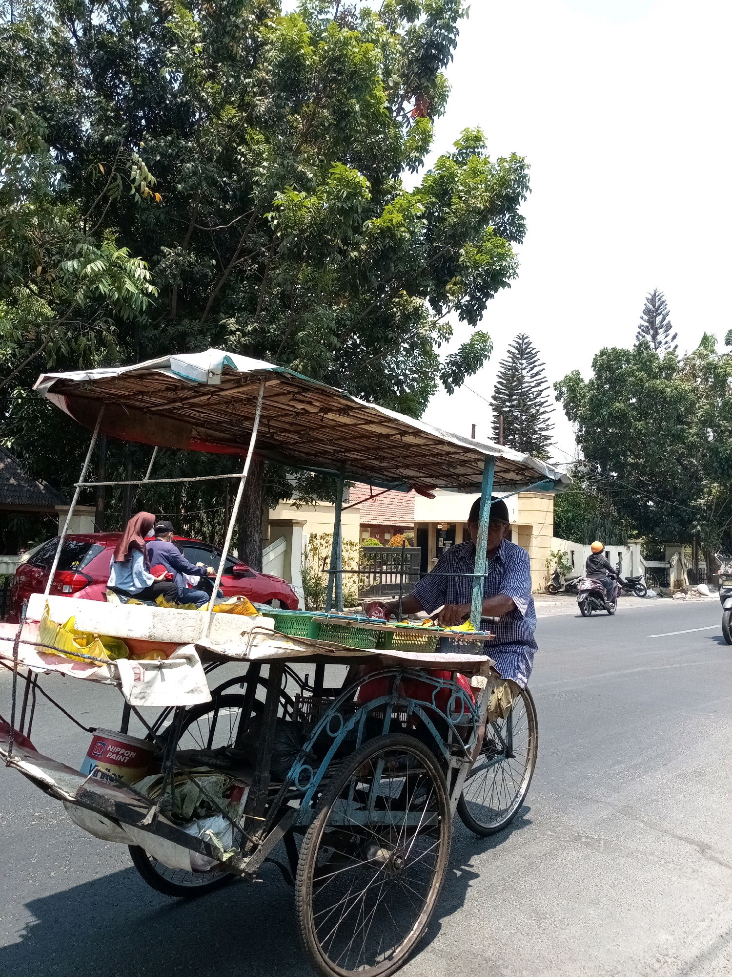 In Bekasi, Indonesia, on a sunny day, a pedicab driver with a checkered shirt and a blue and white striped outfit pedals a three-wheeled vehicle. The pedicab is loaded with goods, including yellow plastic bags and what appear to be food items displayed in a green basket. A woman wearing a maroon hijab and a light blue shirt, along with a person in a dark jacket, are seated in the passenger area of the pedicab. In the background, a red car is parked, and the scene unfolds along a paved road bordered by trees, houses, and other buildings. Motorcyclists are also visible on the road. The pedicab has a canopy overhead, supported by poles, and its structure is a mix of metal and wood. The overall environment suggests a typical street scene in a city in Indonesia.