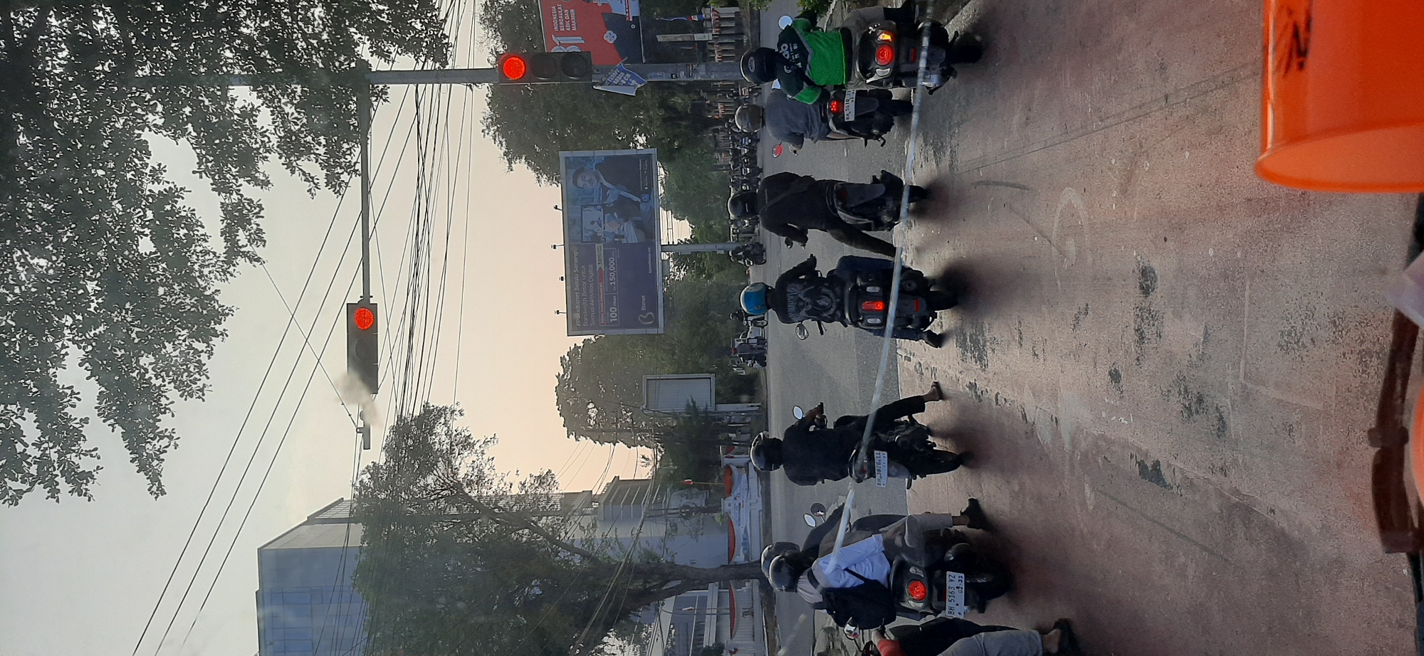 The image captures a street scene in Jambi City, Indonesia, likely during late afternoon or early evening, as indicated by the soft, diffused sunlight and the glowing traffic lights. A line of motorcycles is stopped at a traffic light, suggesting a moment of pause in urban traffic flow. The traffic light overhead is illuminated red, directing the vehicles to halt.
Several individuals are visible on the motorcycles, most wearing helmets, a common safety practice. Their attire varies, with some in casual clothing and one rider in a green jacket, possibly a delivery service uniform. The motorcycles themselves are modern scooters, typical for urban transportation in Indonesia.
In the background, a tall, modern building with reflective glass windows stands prominently, hinting at a commercial or administrative area. Adjacent to it is a large billboard advertising "Biznet," a telecommunications company, displaying pricing and images related to their services. Trees line the street, their branches partially obscuring the sky, and overhead power lines crisscross the scene, indicative of a developed urban environment. The road surface appears to be paved, with some wear and tear and occasional markings, and a red lane divider is visible in the foreground. A portion of an orange object, possibly a traffic cone or container, is present in the very bottom of the frame, suggesting the vantage point is from a vehicle or a similar low perspective. The overall atmosphere is that of a typical busy intersection in a Southeast Asian city, with the waiting traffic and surrounding urban infrastructure.