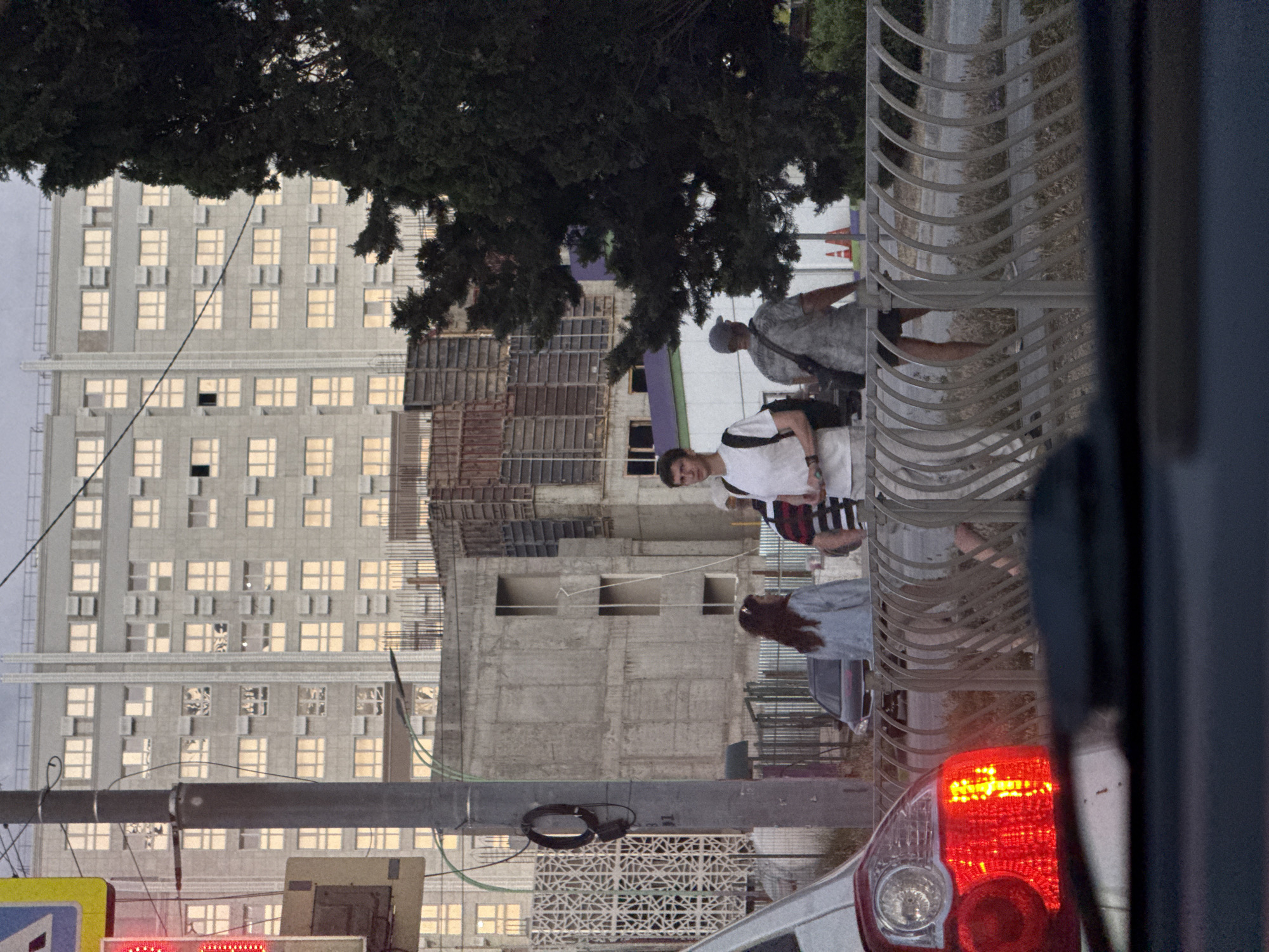 In an urban setting, an array of people are captured in motion, with several individuals positioned near a street and what appears to be construction. In the foreground, a red car taillight is visible, suggesting a vehicle. Behind it, a metal fence with a curved design separates the street from a sidewalk area.
On the sidewalk, a young man in a white t-shirt and shorts is walking, carrying a backpack. He is looking towards the right of the frame. To his left, a woman with dark hair, wearing a light blue denim jacket, is walking with her back to the camera. Another person, wearing a striped t-shirt and a hat, is walking away from the camera, towards the right. In the background, a large, multi-story building dominates the skyline, with many windows illuminated from within. Adjacent to this building, a construction site is evident, with scaffolding and unfinished concrete structures. To the right of the image, a dense green tree stands tall. The overall scene depicts a typical urban environment with a mix of residential buildings, ongoing construction, and pedestrian activity.