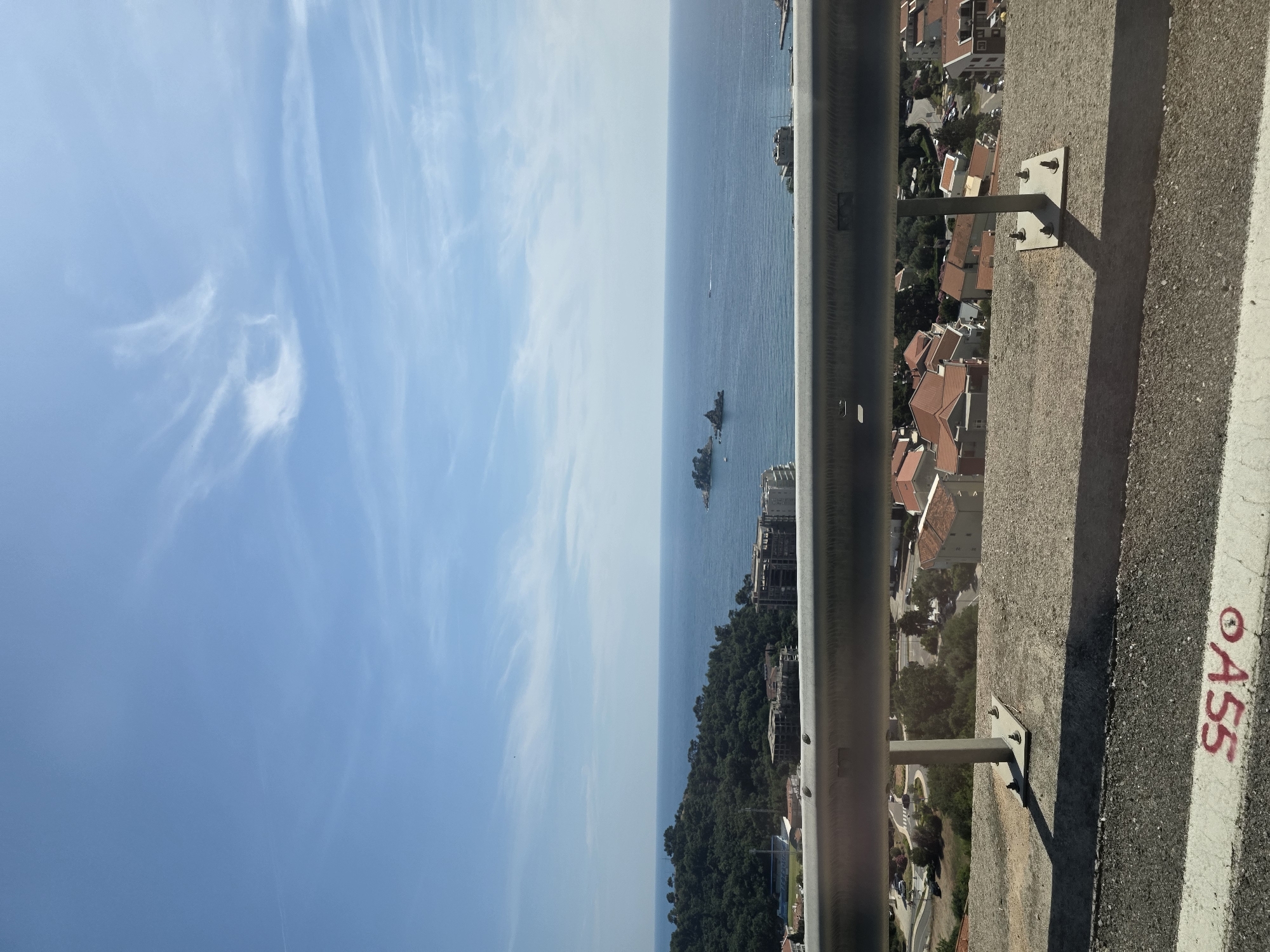 The image captures a panoramic view of the Adriatic Sea from a high vantage point, likely a road overlooking the coastal town of Brežine, Montenegro. The foreground features a weathered concrete barrier with a silver metal guardrail bolted securely onto it. The guardrail casts a distinct shadow on the concrete below. The road surface in the immediate foreground shows signs of wear and tear, with white and red markings visible, including the numbers "ASS".
Beyond the barrier, a densely forested hillside slopes down towards the sea. Nestled among the trees and on the lower slopes are numerous residential buildings with distinctive red-tiled roofs, characteristic of Mediterranean architecture. A few larger, more modern buildings are also present.
The sea stretches out to the horizon, appearing calm and a deep blue. A few small rocky islands are visible in the distance, with a small vessel positioned near them. The sky is a clear, bright blue, with wisps of white cirrus clouds streaking across it. The overall impression is one of a sunny day, with the coastline and sea bathed in natural light. No people are visible in the frame.