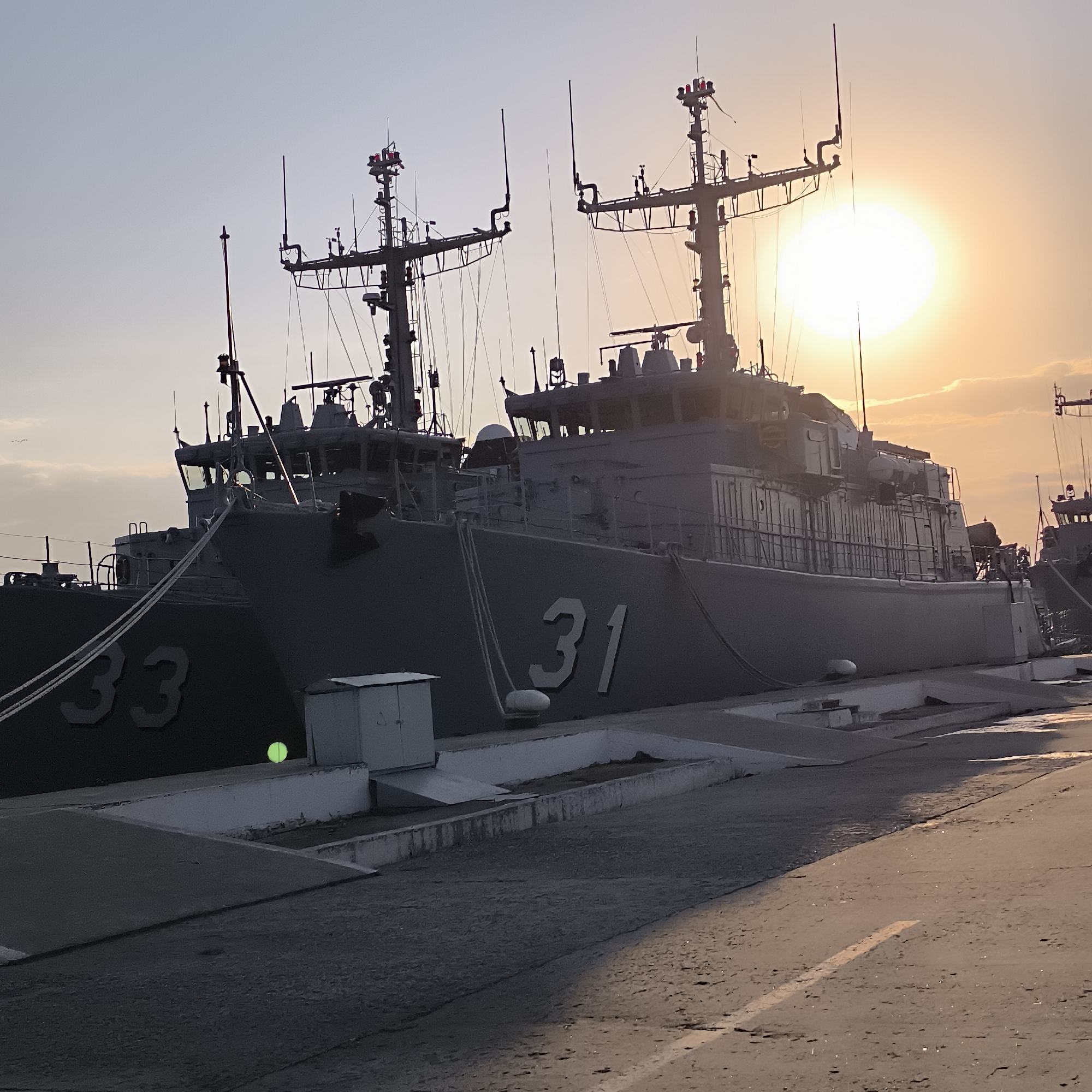The image captures a scene at sunset in Varna, Bulgaria, featuring two naval vessels docked side-by-side. The vessel on the right, identified by the number "31" prominently displayed on its hull, is the primary focus. Its superstructure is detailed with antennas, radar equipment, and navigation lights, silhouetted against the bright, setting sun. To the left, another vessel, marked with "33," is partially visible. Both ships appear to be moored at a dock or pier, indicated by the concrete edge and mooring lines. The ground in the foreground is paved with asphalt, showing some wear and reflecting the warm light of the sunset, with a single yellow line marking a lane. A small, light green spherical object rests on the edge of the dock near the "33" vessel. The sky is a gradient of pale blue and warm orange hues, with a few wispy clouds near the horizon. The overall atmosphere is serene, characteristic of a calm evening by the sea.