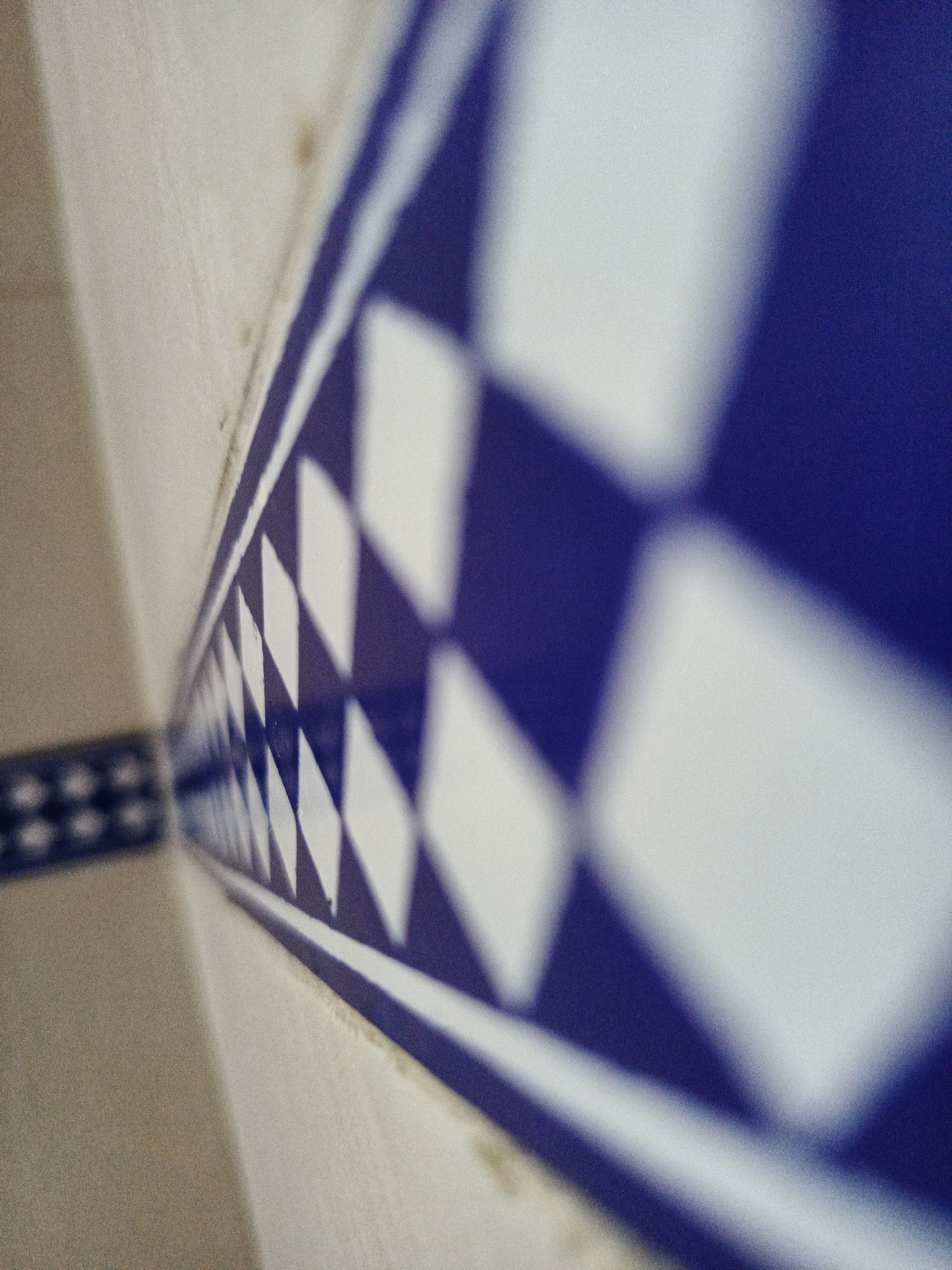 A close-up, slightly out-of-focus image shows a blue and white patterned tile border running along a wall. The tiles alternate between blue and white diamonds. The wall adjacent to the tile border appears to be beige or cream-colored. The background is out of focus, but suggests more tile work with a small square pattern. The image does not provide enough information to determine the precise location in Los Robles, Venezuela. There is no information about people, actions or interactions visible in the image.