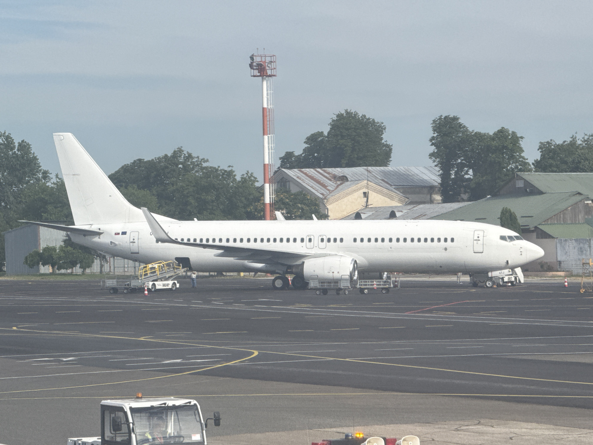 A white passenger airplane is parked on an airport tarmac under a hazy sky. The airplane is a Boeing 737, identified by its distinctive engine nacelles and wing shape. A mobile staircase is positioned next to the front passenger door, and ground crew are visible near the aircraft. In the background, several airport buildings and a red and white striped communications tower are present. The tarmac is marked with various yellow and white lines, indicating taxiways and parking positions. In the foreground, the front of a white tug vehicle is visible. The overall scene suggests a typical airport environment with aircraft and ground operations. The presence of a Boeing 737, along with the airport infrastructure, points to Chișinău International Airport in Moldova as the likely location.