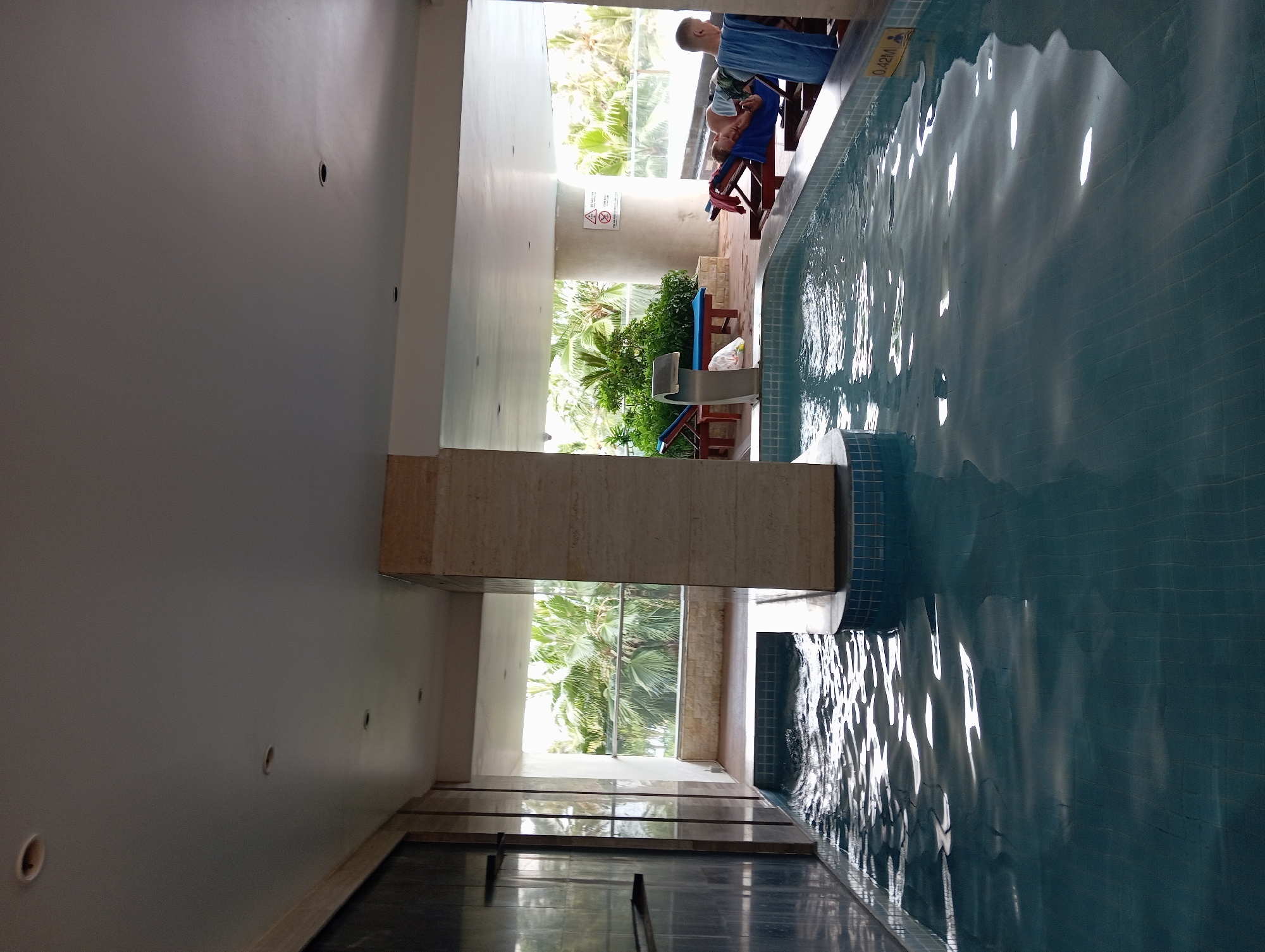 The image shows an indoor swimming pool area at a resort in Nha Trang, Vietnam. In the foreground, a body of water with rippling surface reflects the overhead lighting and is surrounded by tiled edges. A central column, clad in beige stone tiles, rises from the pool area. Beyond the pool, there is an open-air section with wooden decking. Several lounge chairs are placed on the decking, with two men relaxing on them. One man, wearing a dark top and shorts, is lying on a chair with a blue towel draped over him. The other man, seen from the side, is wearing a patterned shirt and appears to be looking out. In the background, lush green palm trees and other vegetation are visible through a railing, suggesting a tropical outdoor environment. The overall scene depicts a tranquil and luxurious resort setting.