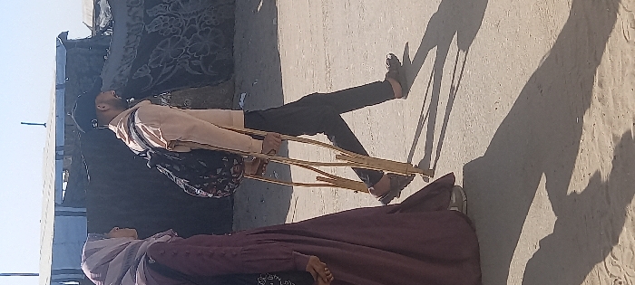 The image shows two individuals walking on sandy ground under bright daylight in Khan Yunis, Palestinian Territory. On the left, a female wears a maroon abaya and a matching hijab, her face in profile looking forward. On the right, a male figure with a beard wears a dark baseball cap, a light beige long-sleeved shirt, dark trousers, and dark slip-on sandals. He carries a dark, patterned backpack and uses two wooden crutches for mobility. Both are in motion, walking from right to left across the frame. Long, distinct shadows of the individuals and crutches are cast on the ground to their left. The background features makeshift structures constructed from light-colored and dark patterned tarpaulins or fabrics.