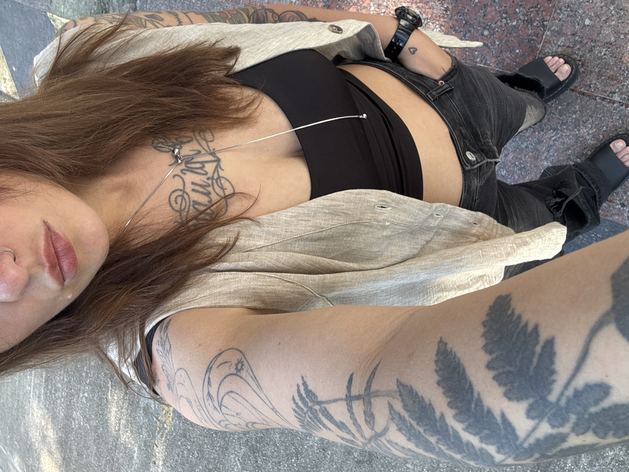 The image displays a high-angle, waist-up selfie of an individual in an outdoor urban setting within the city of Sochi, Russia. The subject, appearing to be female, has long, straight brown hair. A silver nose ring is present on the left nostril.
The individual is dressed in a light-colored, possibly beige or off-white, sleeveless vest worn open over a black crop top. Dark grey or black distressed jeans are worn on the lower body. Black open-toe sandals are visible on both feet.
Prominent tattoos are visible on both arms and the chest. The left arm features a large, dark fern-like leaf pattern extending along the forearm and upper arm, along with a stylized outline of a heart containing swirls and a starburst design on the upper arm. The right arm displays a full sleeve of dense, dark, abstract or organic patterns. A chest tattoo consisting of black script, appearing to be in Cyrillic, is situated above the cleavage. A small, solid black heart tattoo is located on the back of the right hand, near the wrist.
Accessories include a thin silver chain necklace with a small, stylized pendant and a secondary small silver bead, extending down the chest. A black digital watch is worn on the right wrist. The subject's right hand is placed into the front pocket of their jeans.
The ground surface beneath the individual consists of textured grey asphalt or concrete in the foreground, transitioning to a mottled reddish-brown and lighter-colored granite or stone paving towards the background and right side of the frame. The lighting suggests an outdoor daytime environment.